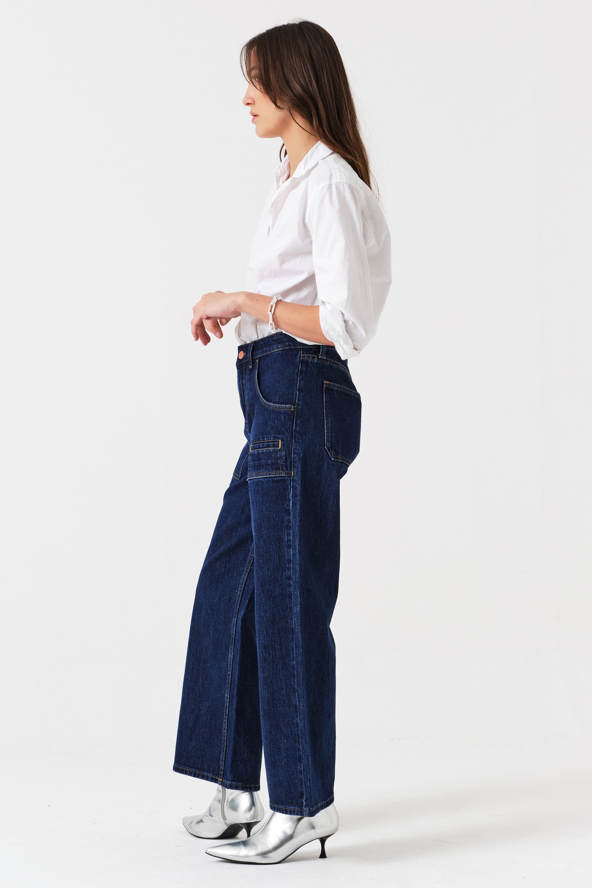 Woman wearing dark wash blue cropped wide-leg jeans with pocket details and a white shirt on a white background, photographed from the side