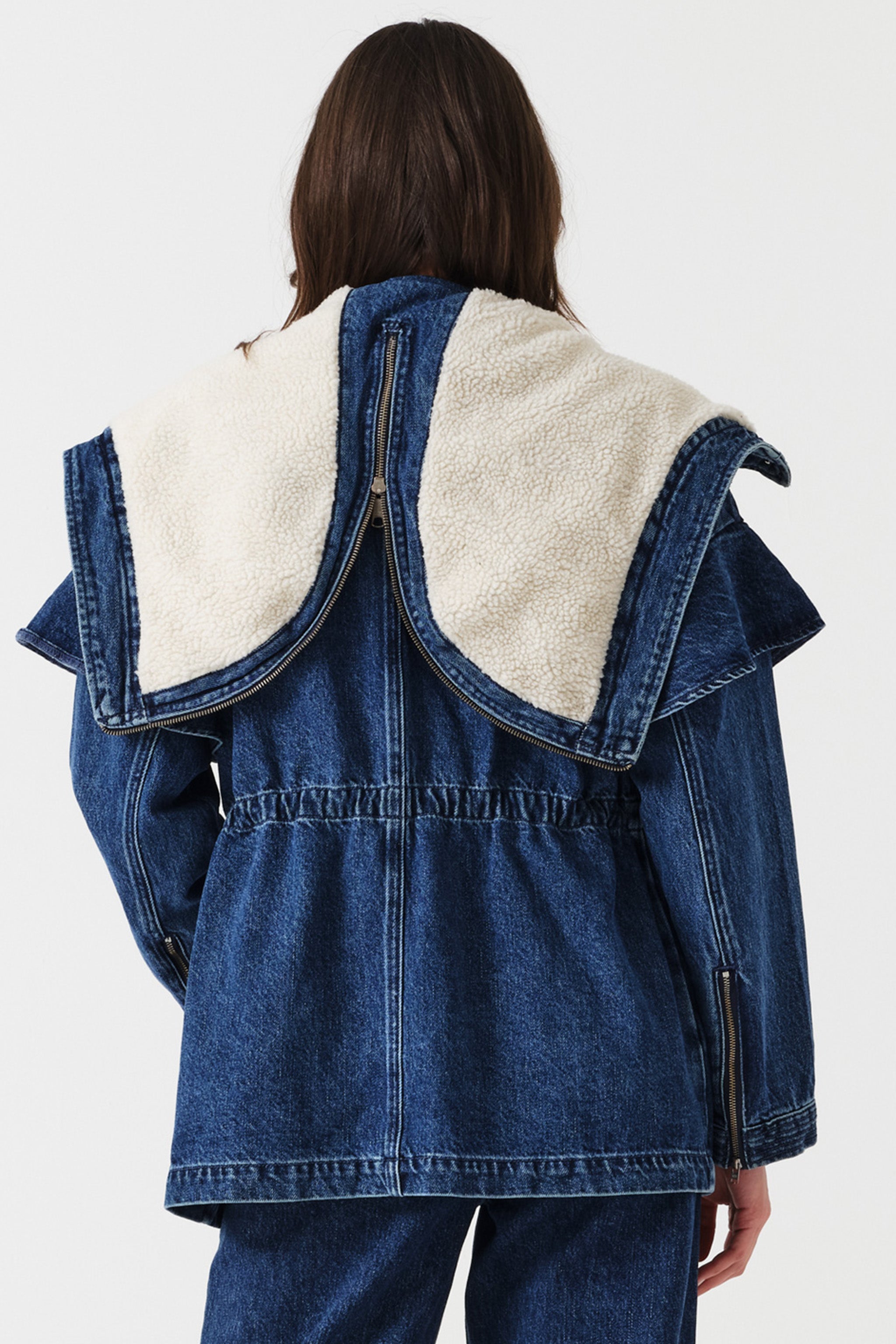 Woman wearing a dark blue denim jacket with a white sherpa lined removable hood, waist cinch, and zip details on a white background, photographed from the back