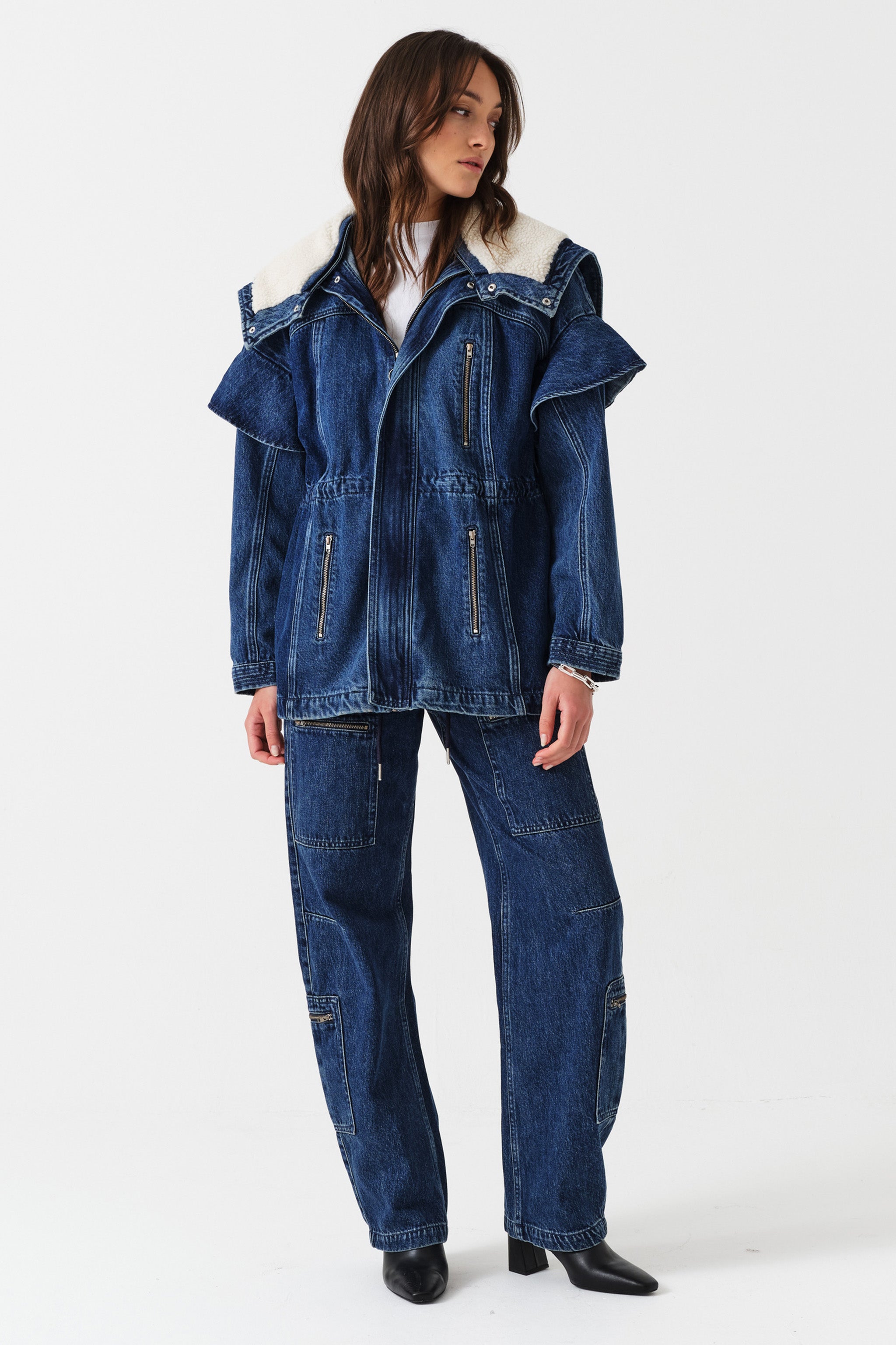 Woman wearing a dark blue denim jacket with a white sherpa lined removable hood, waist cinch, and zip pocket details with dark blue jeans with cargo zip pockets on a white background.