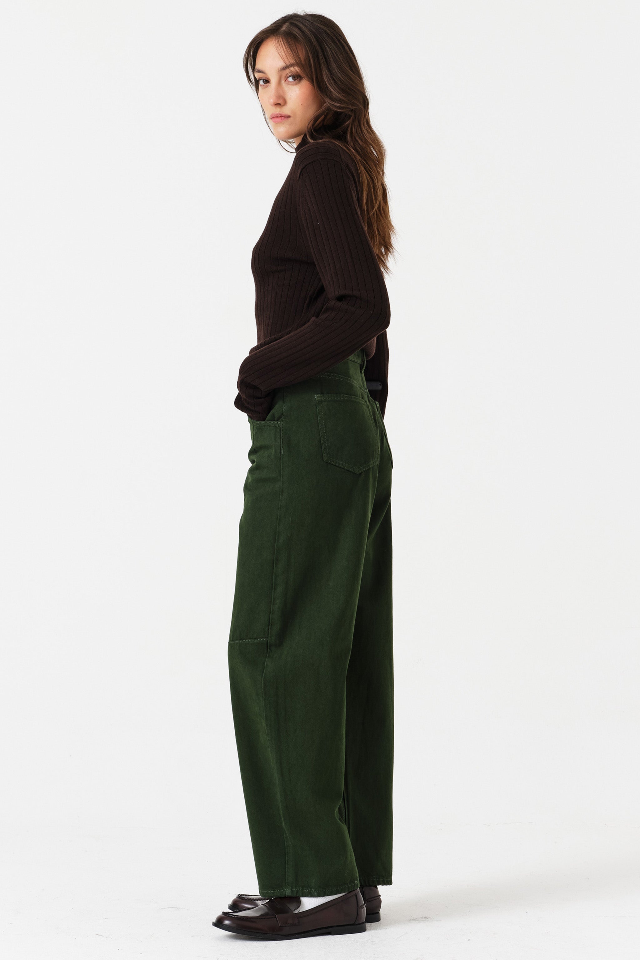 Woman wearing dark green barrel leg jeans and a brown shirt on a white background, photographed from the side