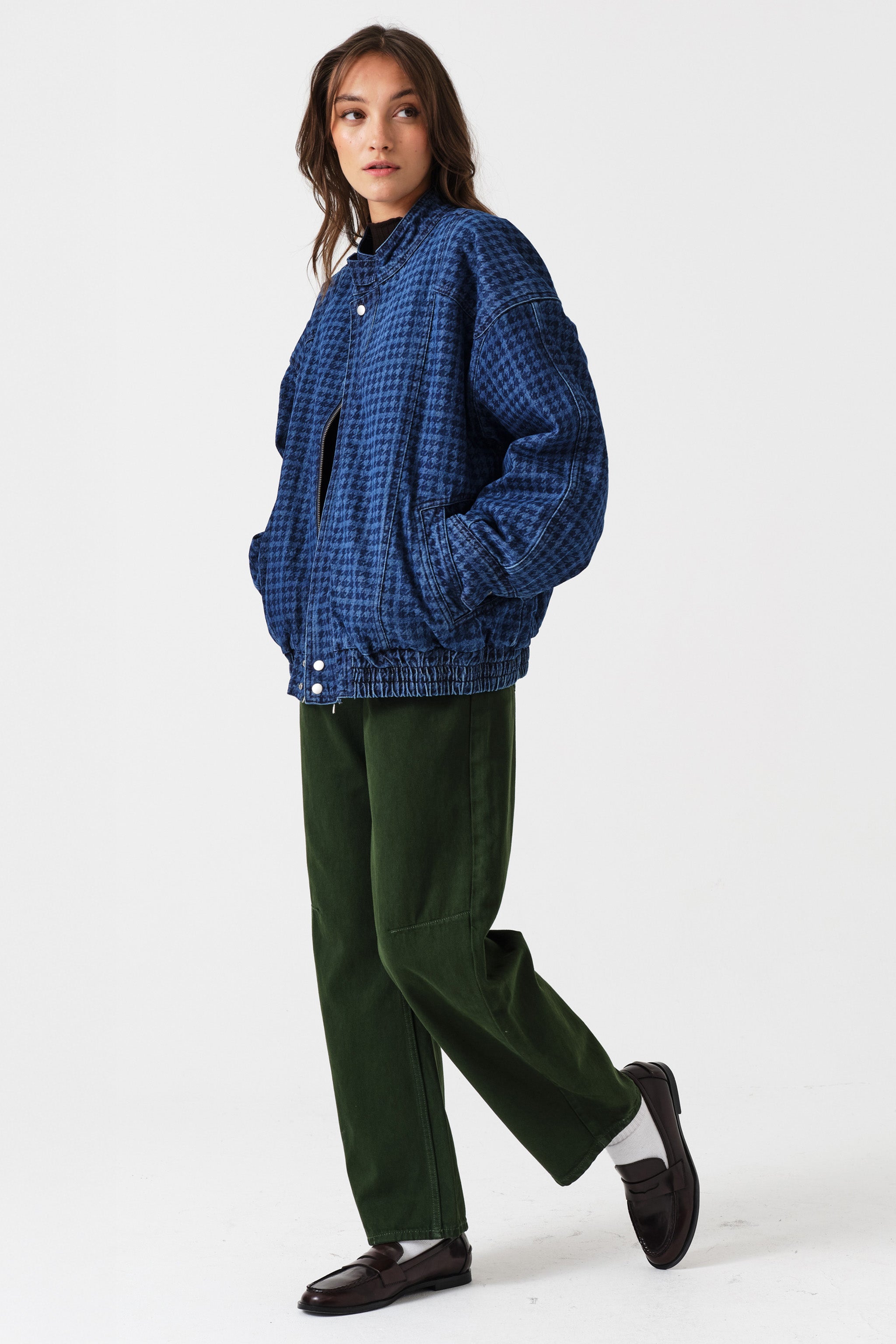 Woman wearing dark green barrel leg jeans, a brown shirt, and a dark blue houndstooth bomber jacket on a white background