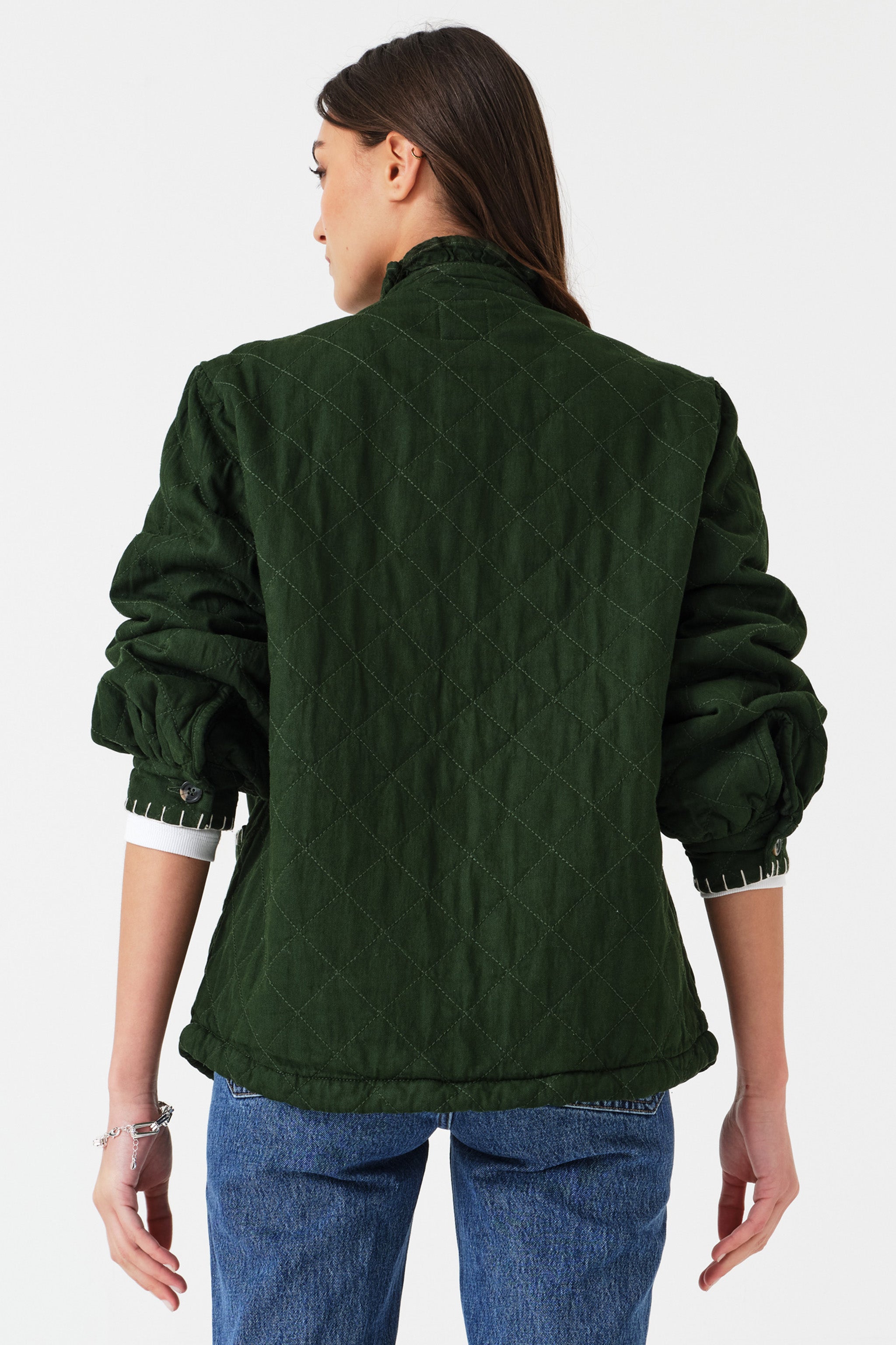 Woman wearing a quilted dark green jacket with white embroidered details and blue jeans on a white background photographed from the back