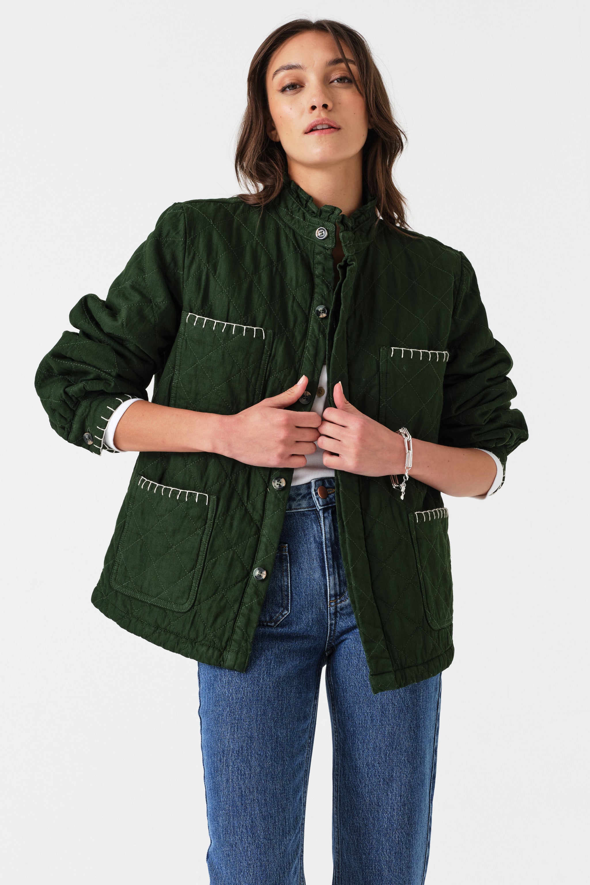 Woman wearing a quilted dark green jacket with white embroidered details and blue jeans on a white background