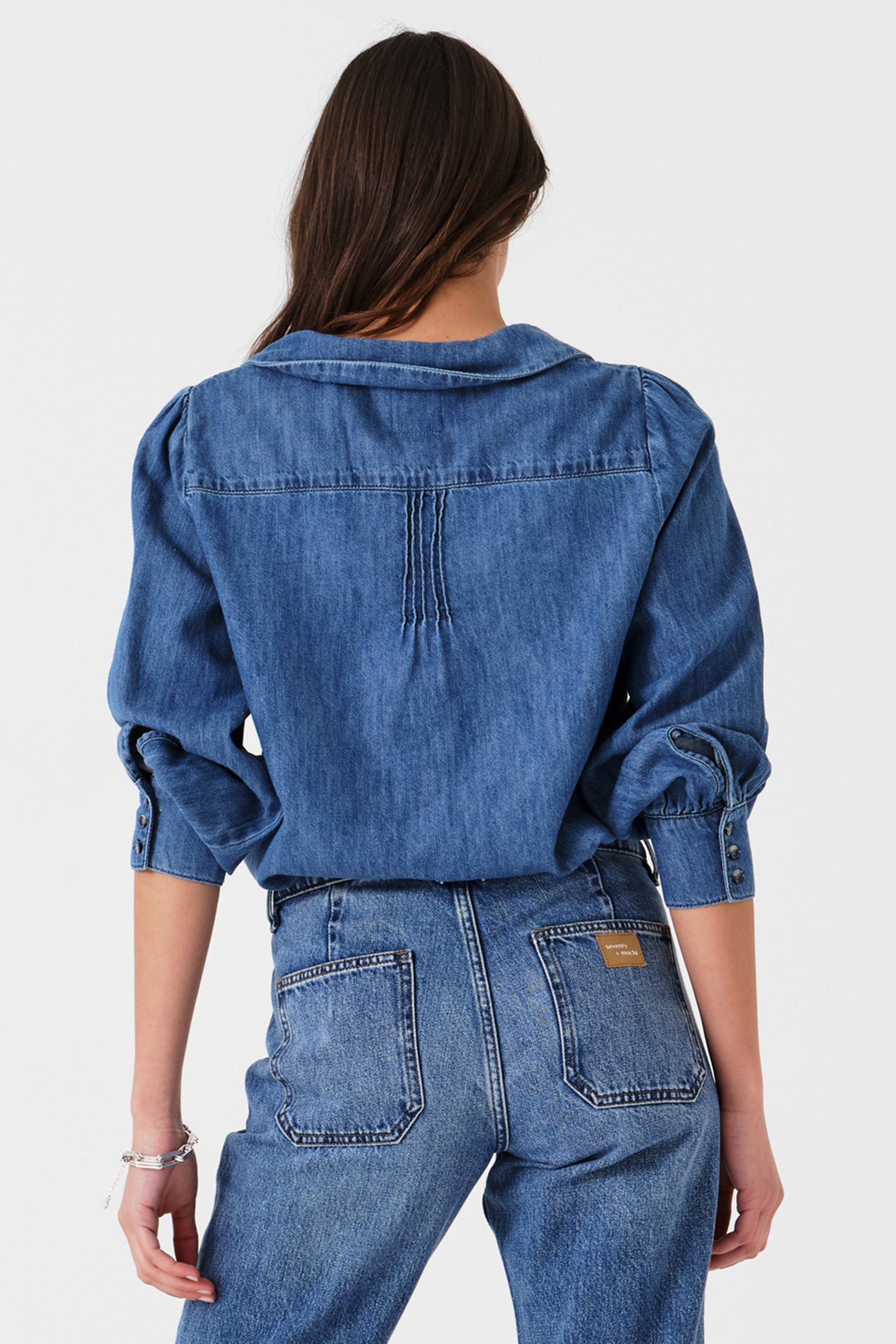 Woman wearing a blue denim blouse with embroidered details and jeans on a white background photographed from the back