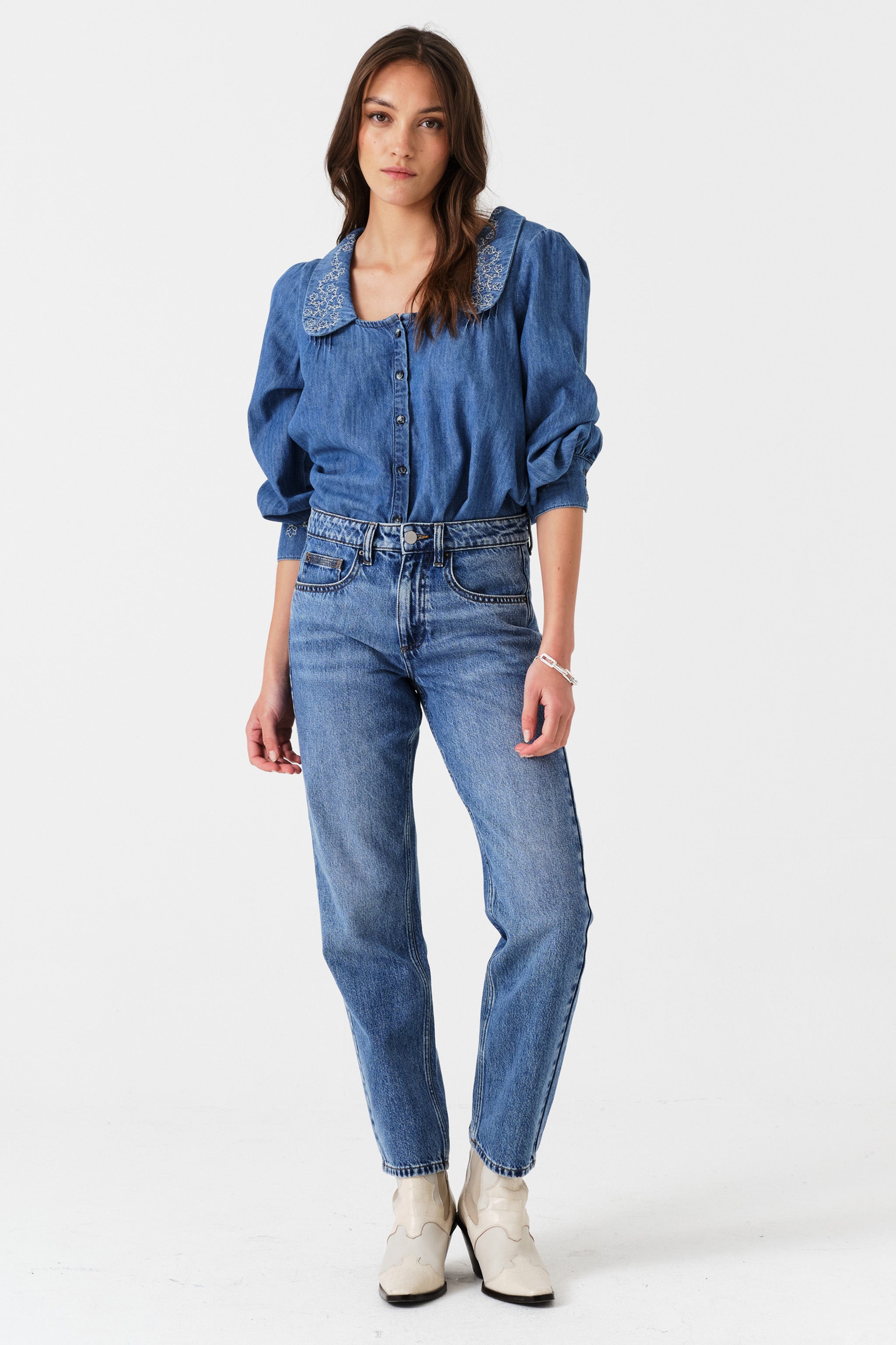 Woman wearing a blue denim blouse with embroidered details and jeans on a white background