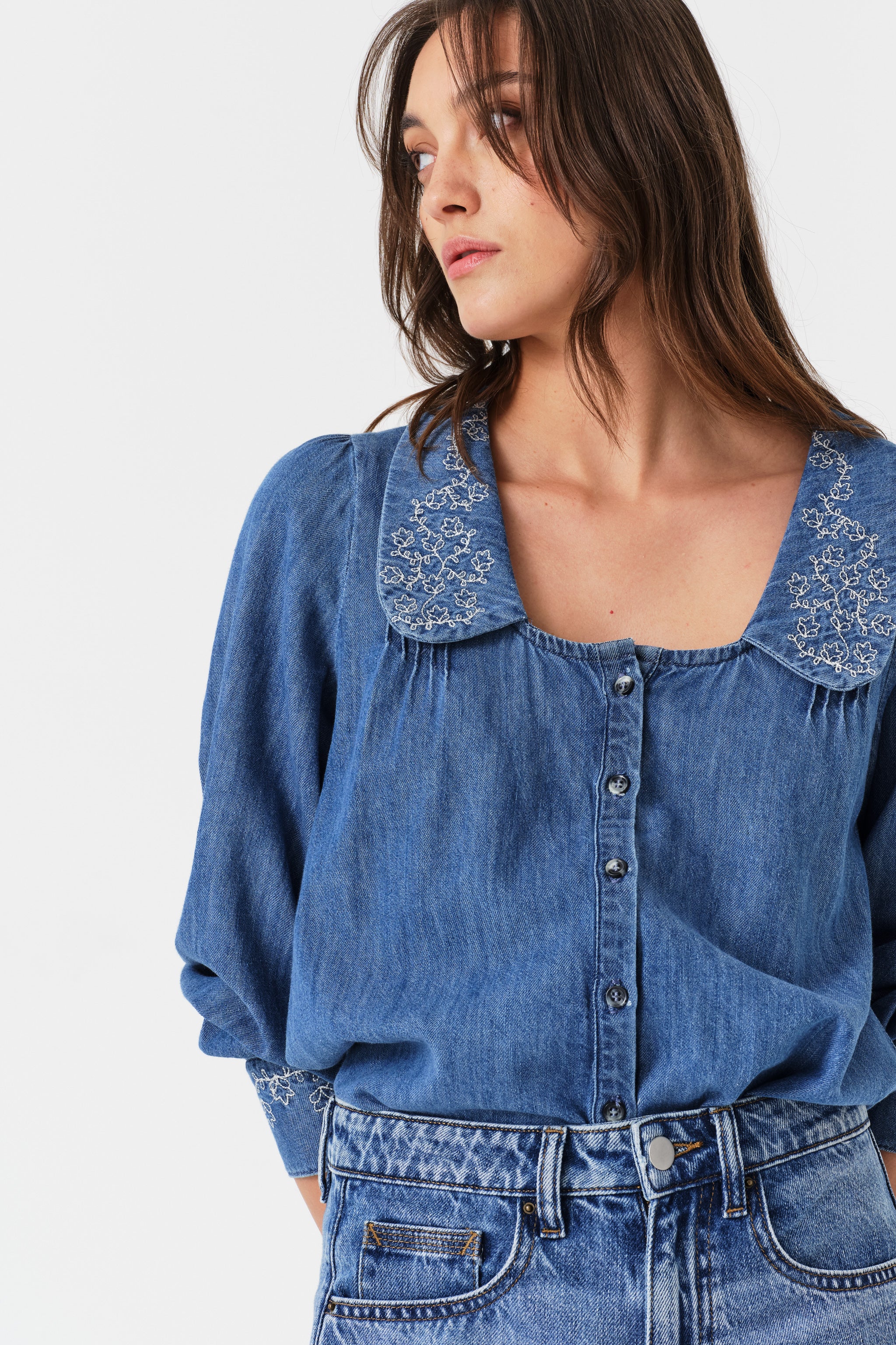 Woman wearing a blue denim blouse with embroidered details and jeans on a white background