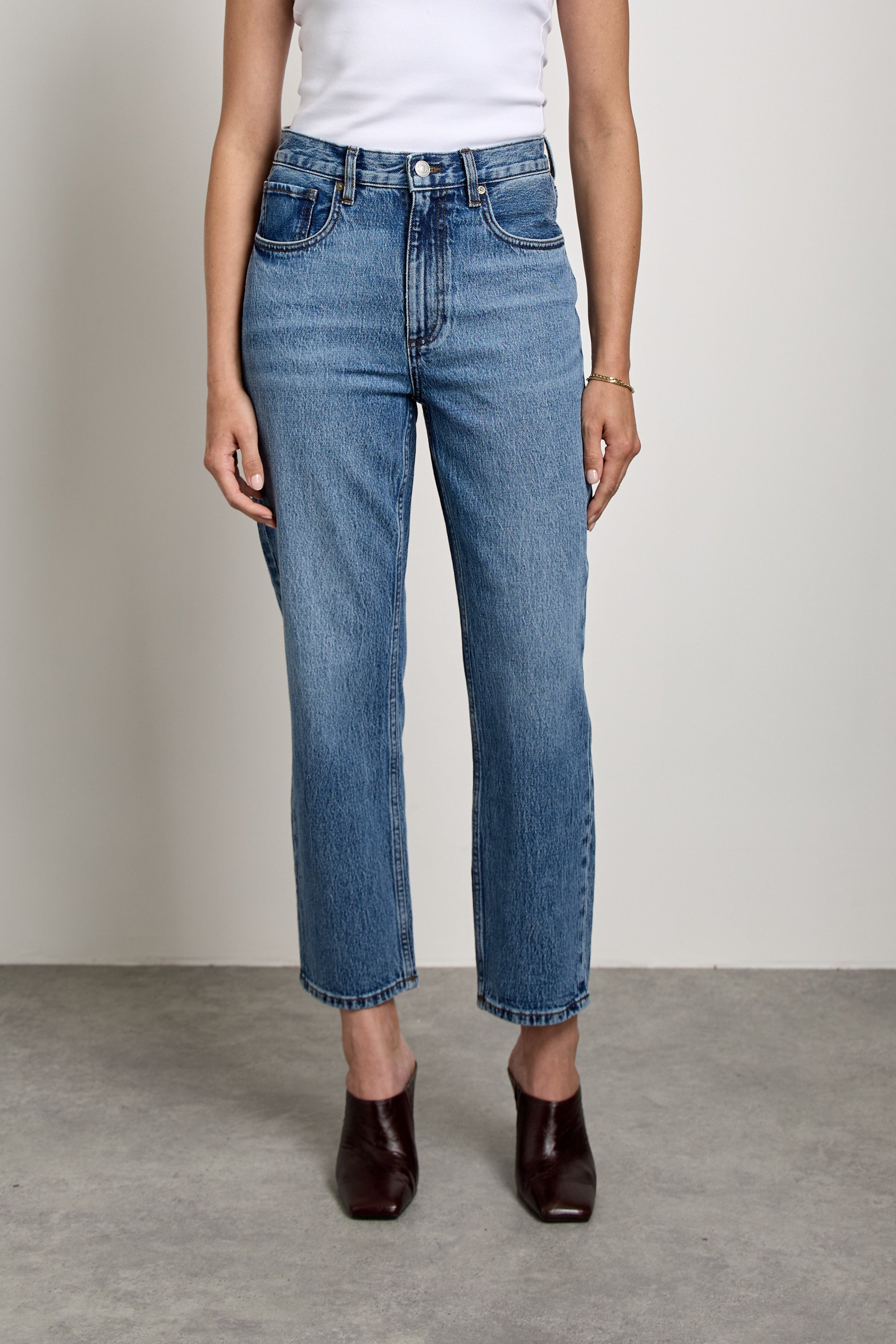 A woman stands in a studio wearing blue cropped jeans, a white tank top, and dark brown shoes against a light gray wall and concrete floor.