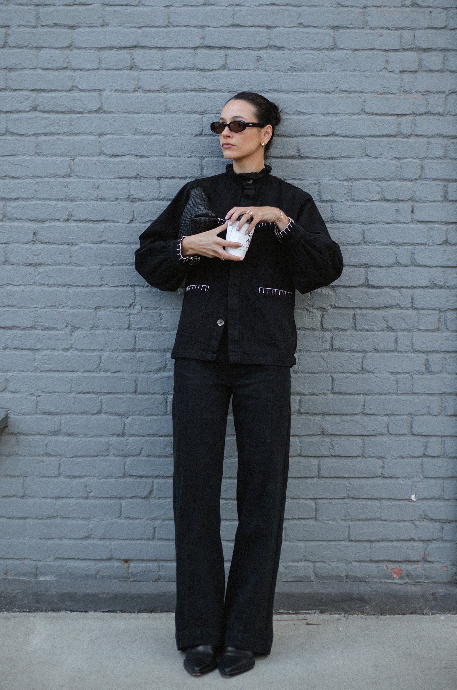 woman holding coffee wearing a denim jacket with white stitching and black high rise, wide leg jeans with a sailor button front fastening detail outside
