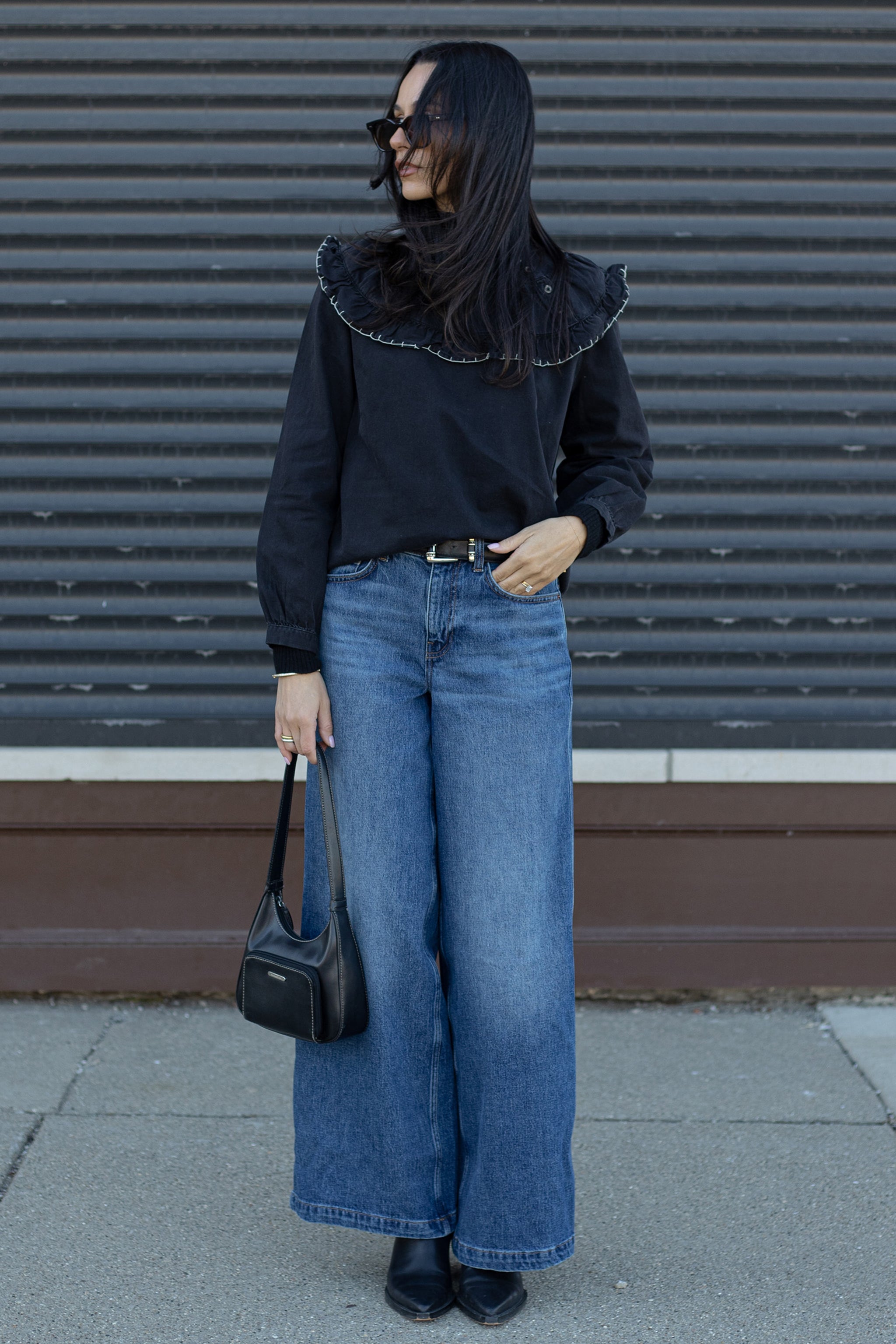 Woman wearing a black shirt with blue wide leg jeans, holding a black bag and standing against a metallic shutter.