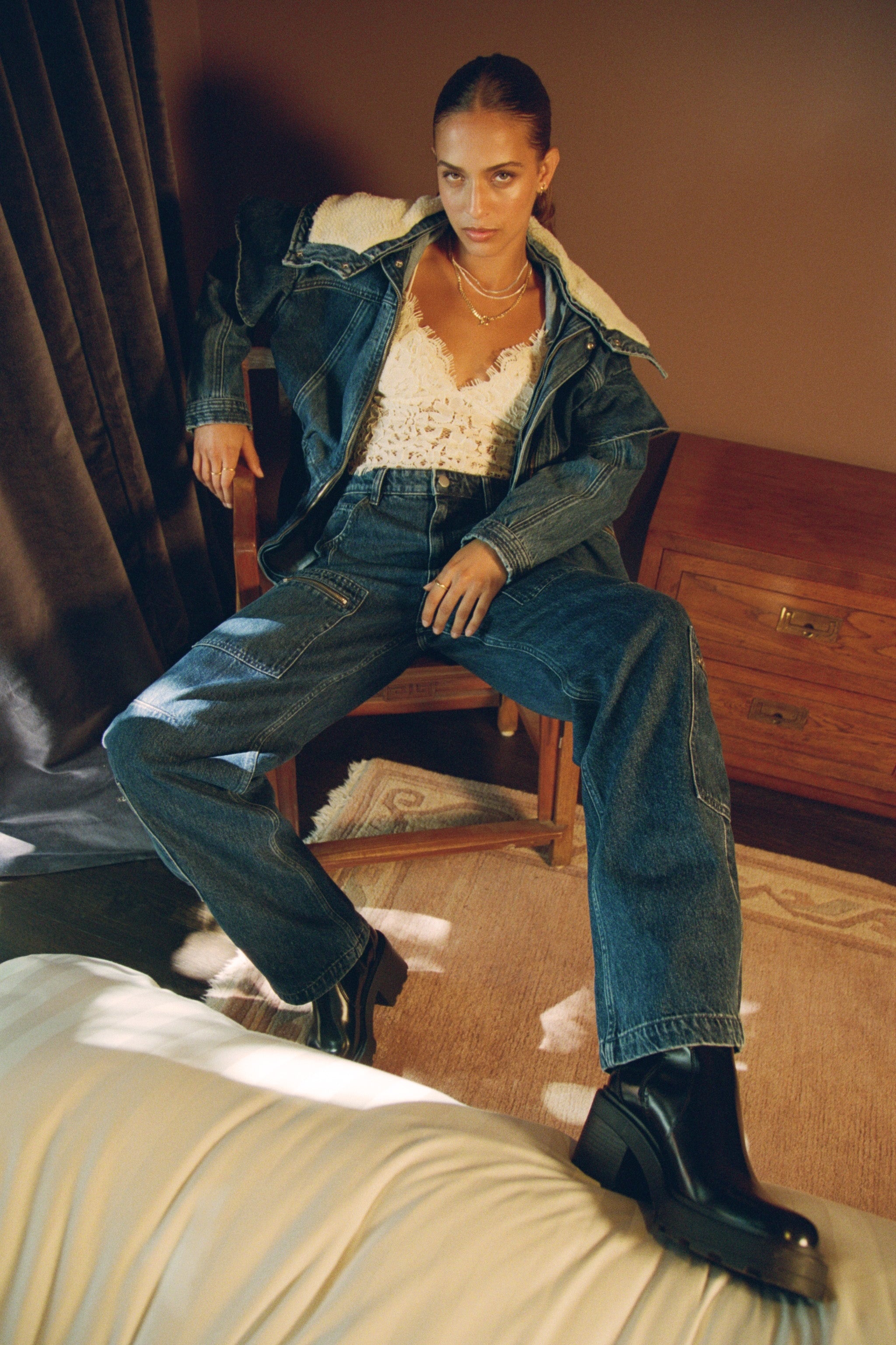 Woman sitting on a chair wearing a blue denim jacket with a sherpa collar and zip details, white top, and blue jeans  with cargo zip pockets in a room.