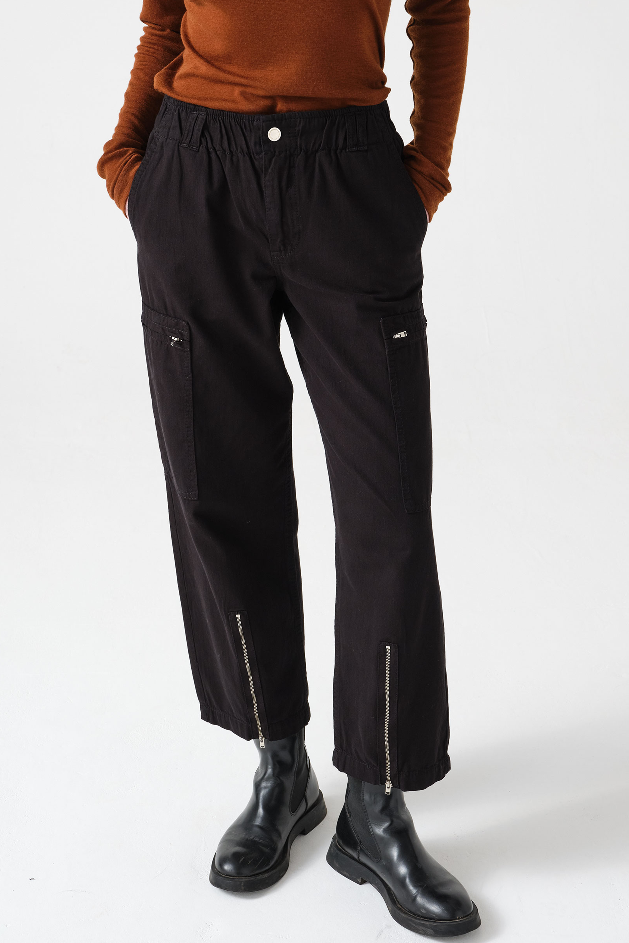 woman wearing a pair of black, lightweight, cargo style pants with an elastic waist, deep zip pockets, and a zip detailed hem paired with an orange top and black boots against a white backdrop