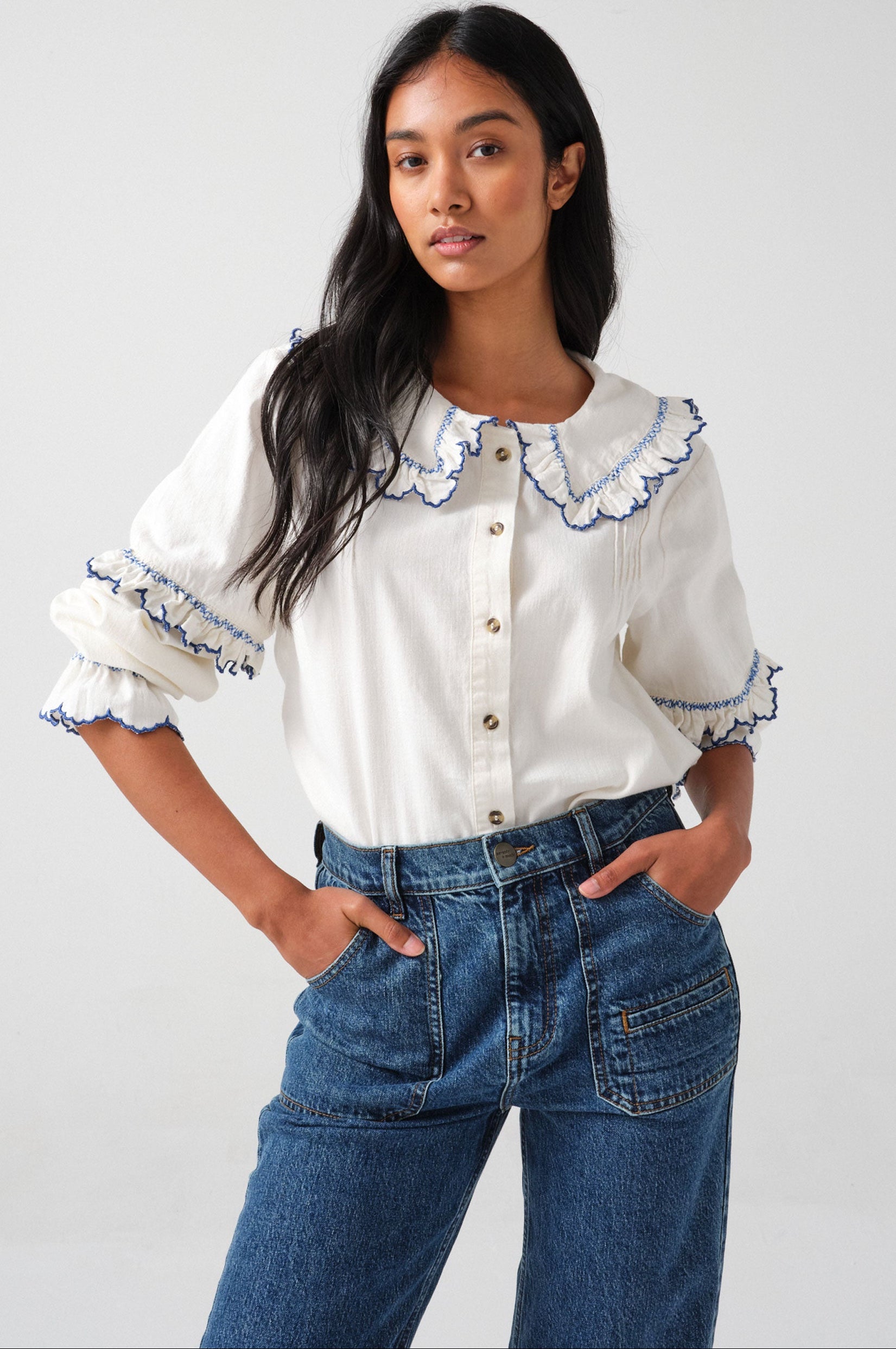 woman wearing an organic cotton  blouse with electric blue frill embroidery on the collar and sleeves, paired with blue jeans.