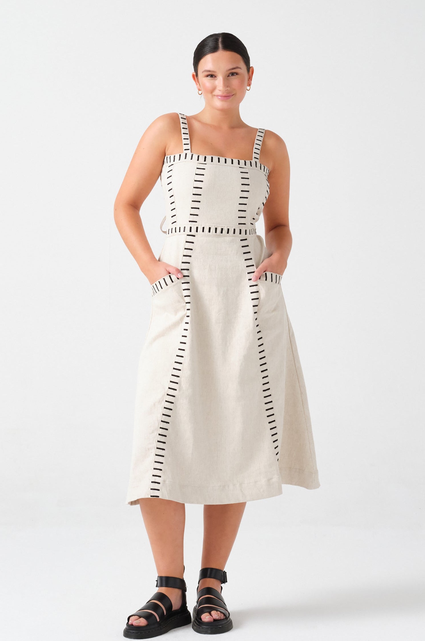 A woman wearing an ecru midi denim dress with black stitch details, a square neckline, front pockets, and an open, lace-up back on a white background