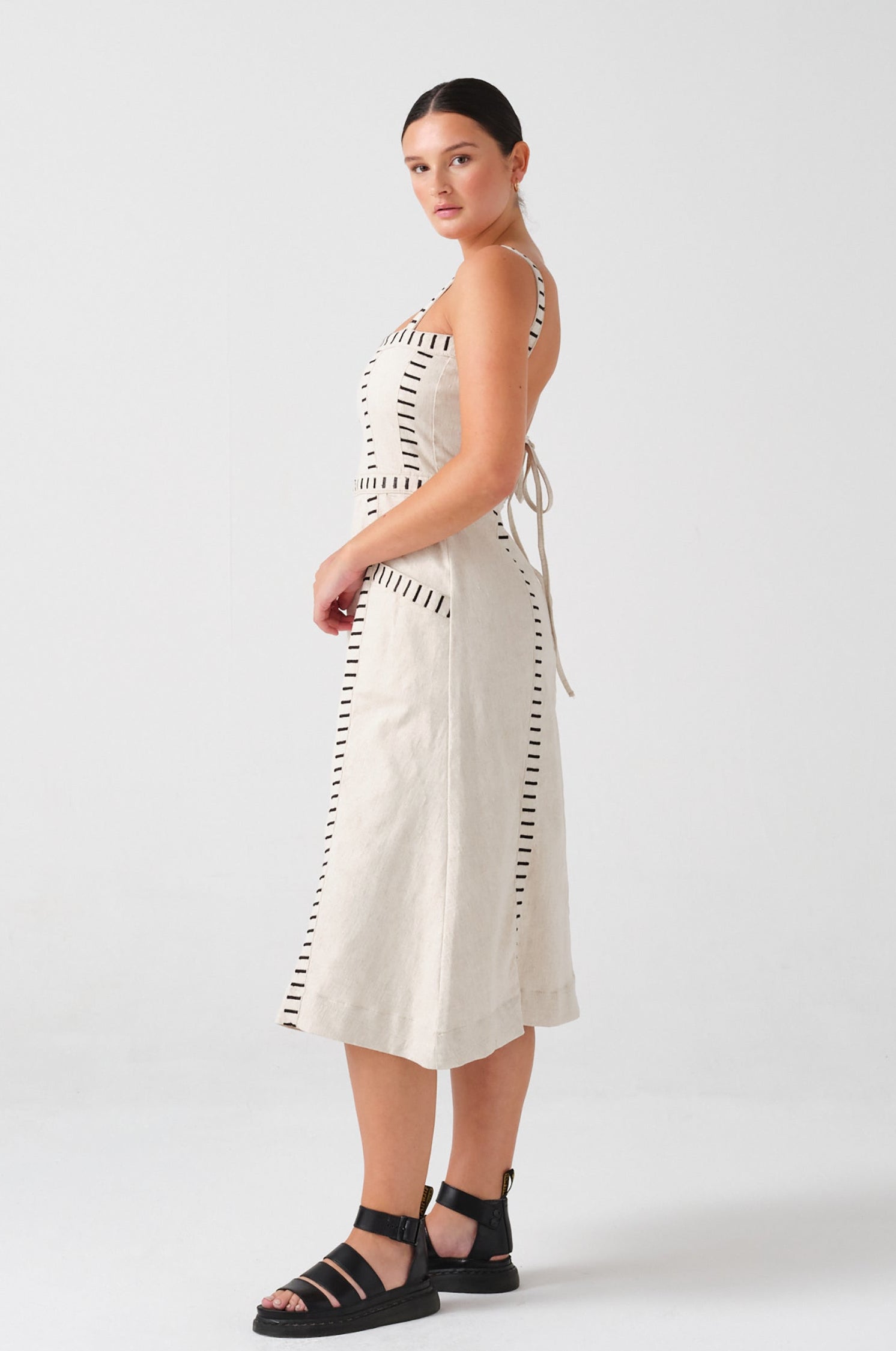 A woman wearing an ecru midi denim dress with black stitch details, a square neckline, front pockets, and an open, lace-up back on a white background