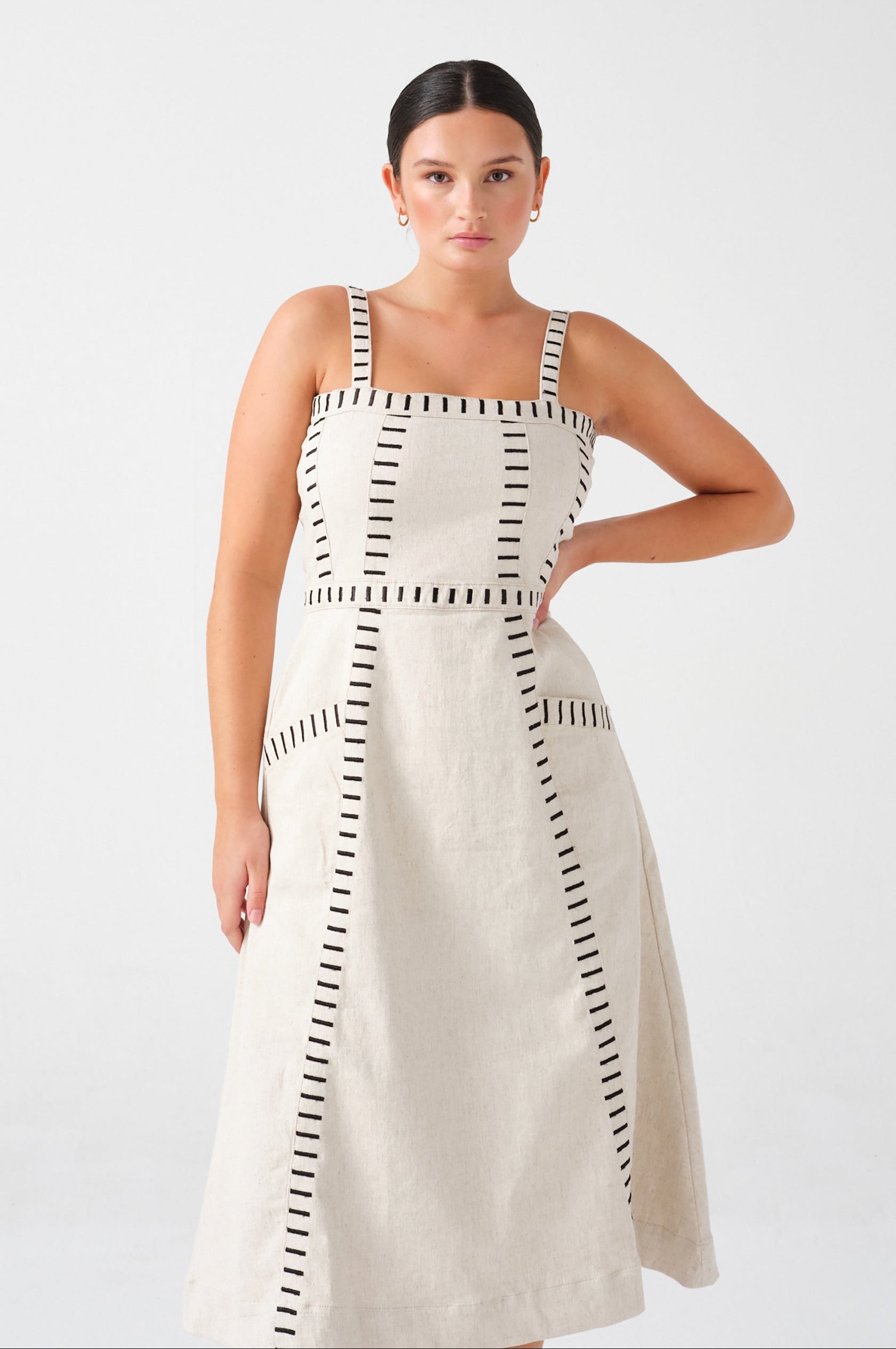 A woman wearing an ecru midi denim dress with black stitch details, a square neckline, front pockets, and an open, lace-up back on a white background