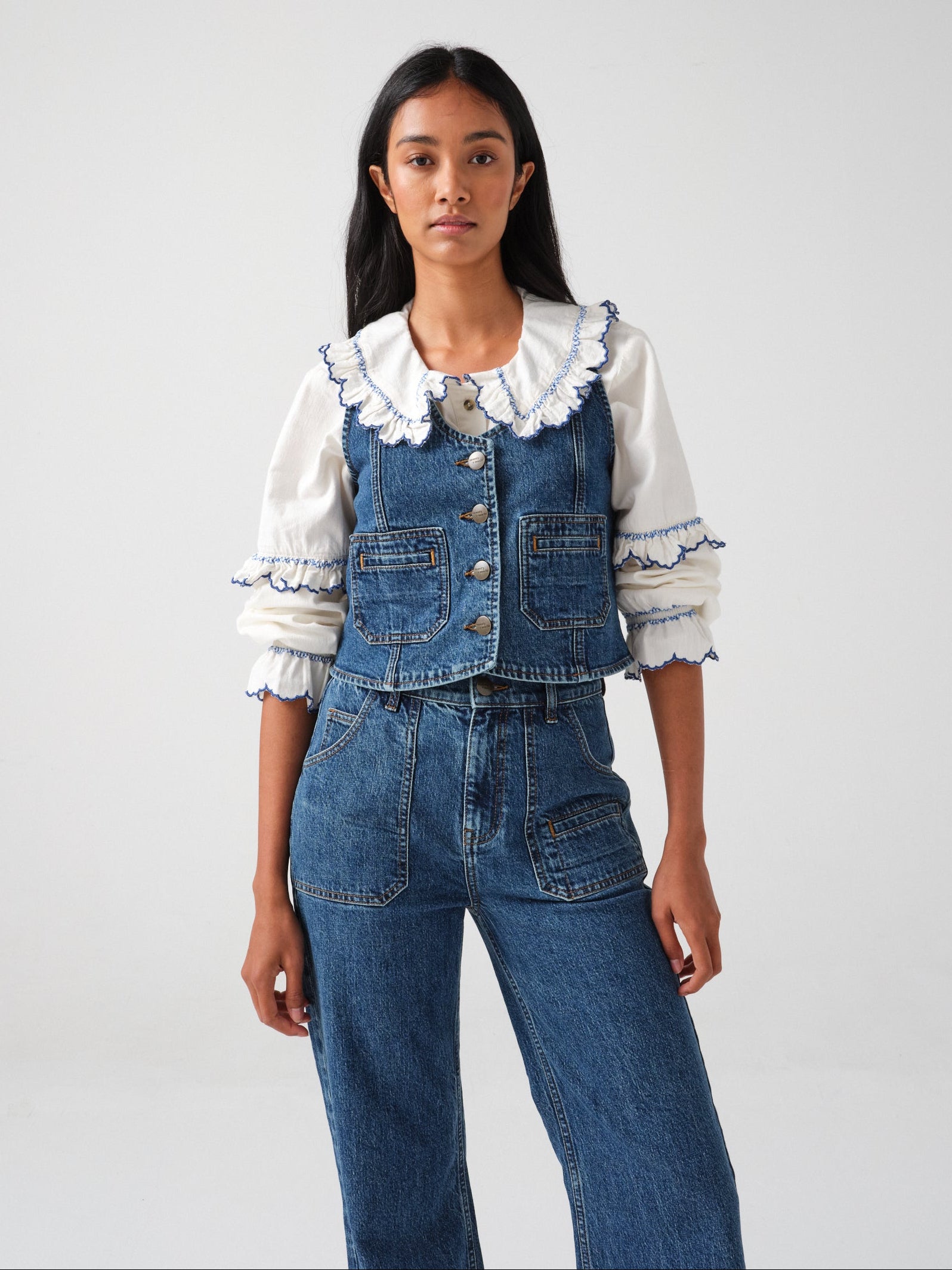 woman wearing an organic cotton  blouse with electric blue frill embroidery on the collar and sleeves, paired with blue jeans and a blue denim vest/ waistcoat