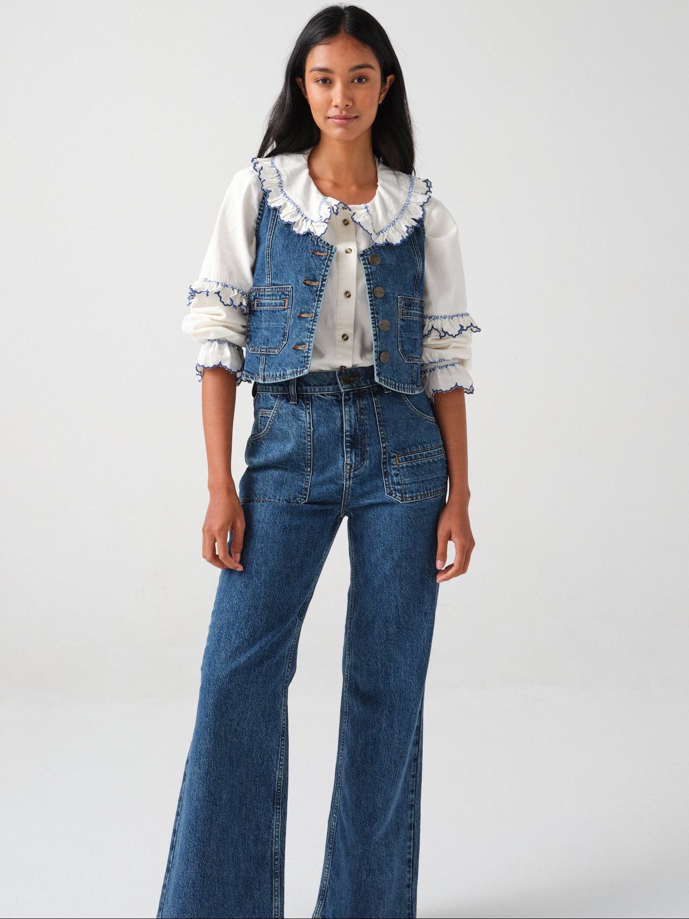 woman wearing an organic cotton  blouse with electric blue frill embroidery on the collar and sleeves, paired with blue jeans and a blue denim vest/ waistcoat