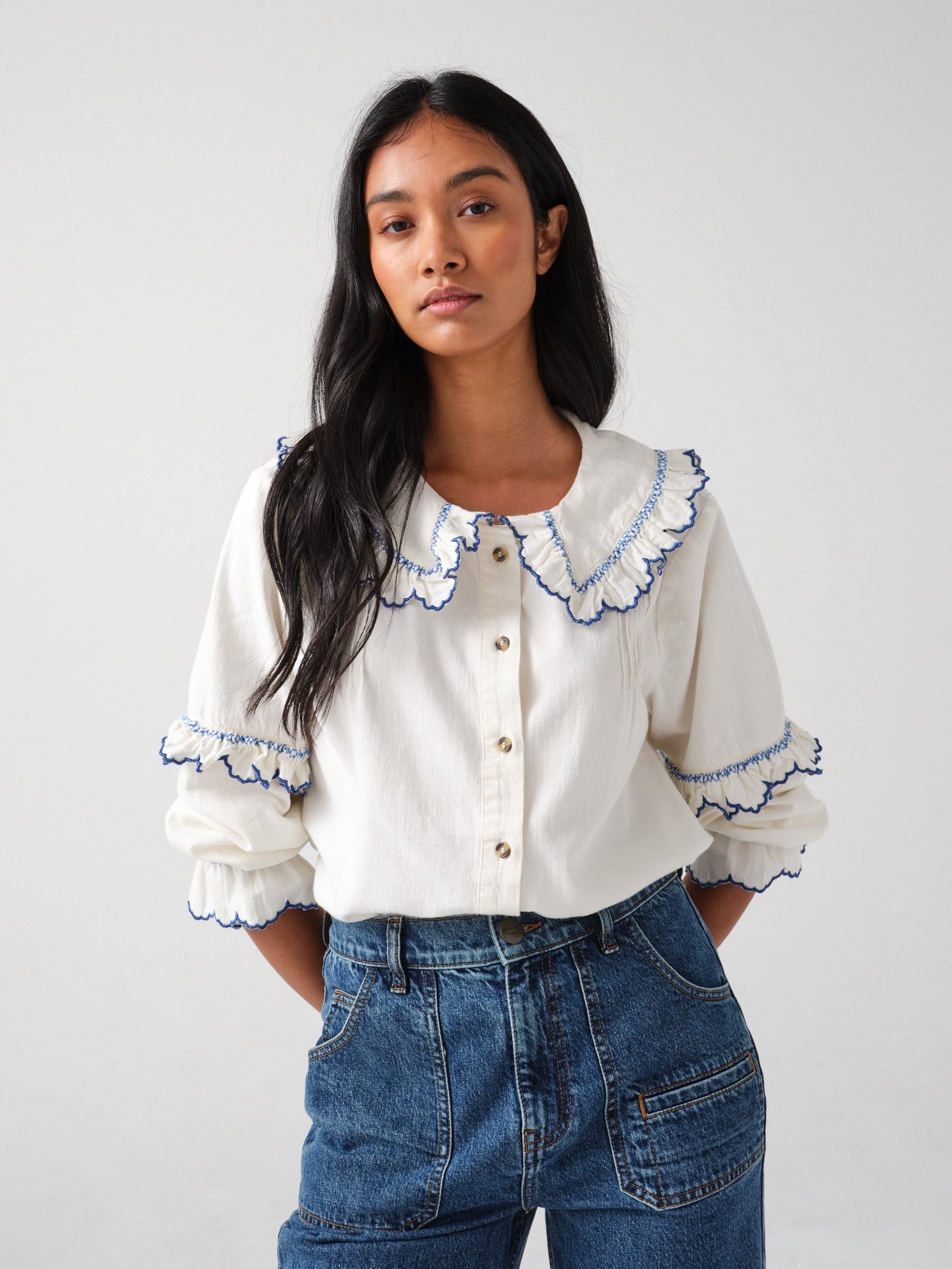 woman wearing an organic cotton  blouse with electric blue frill embroidery on the collar and sleeves, paired with blue jeans on a white background