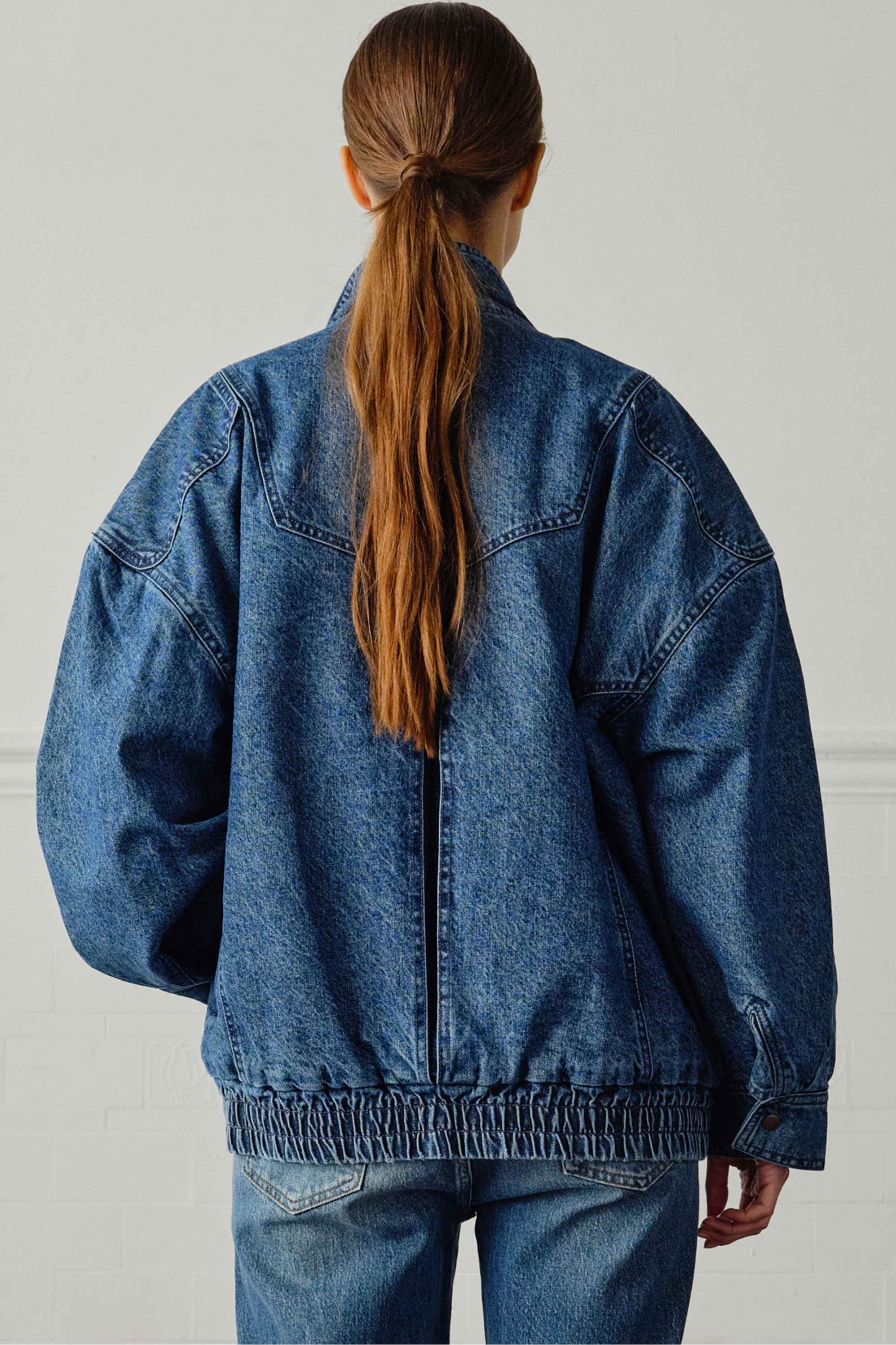 A woman wearing an oversized padded and seamed blue denim retro style bomber jacket with a round neckline, zip, and button closures at the top, bottom, and collar with a white top and blue jeans against a white backdrop