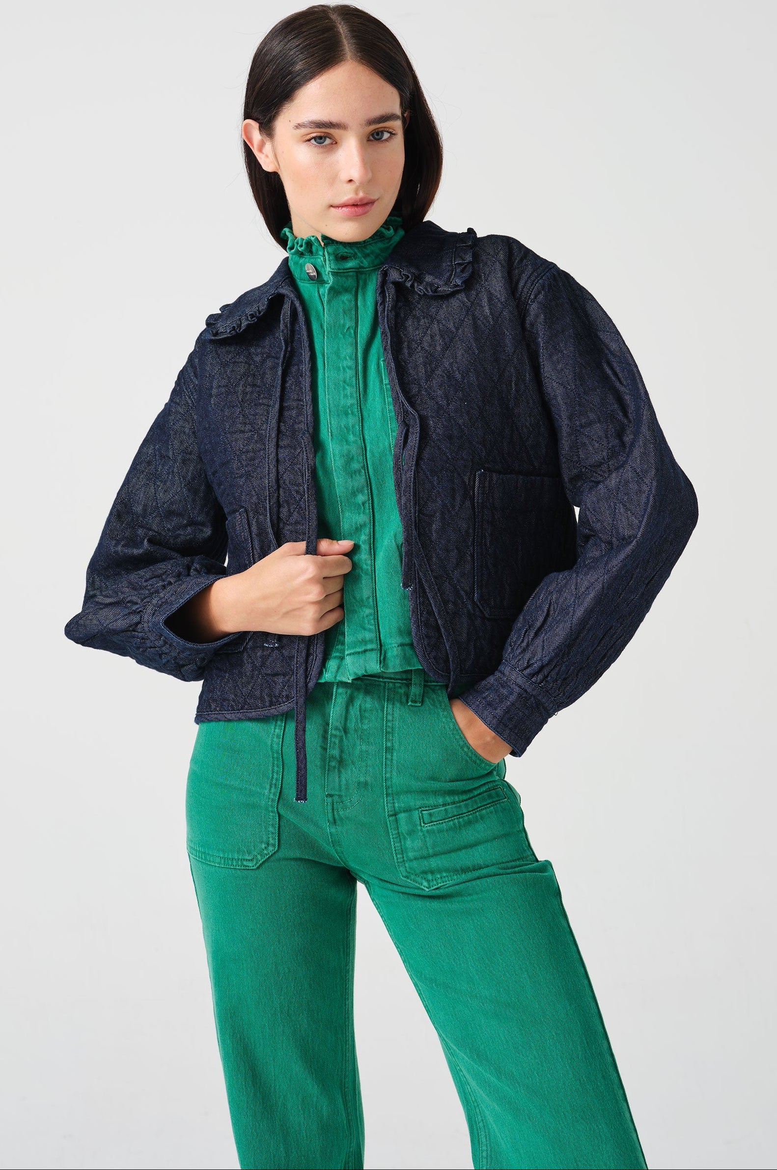 Woman wearing a dark blue denim quilted jacket with a slightly frilled collar and tie closures with a green denim shirt and green denim jeans