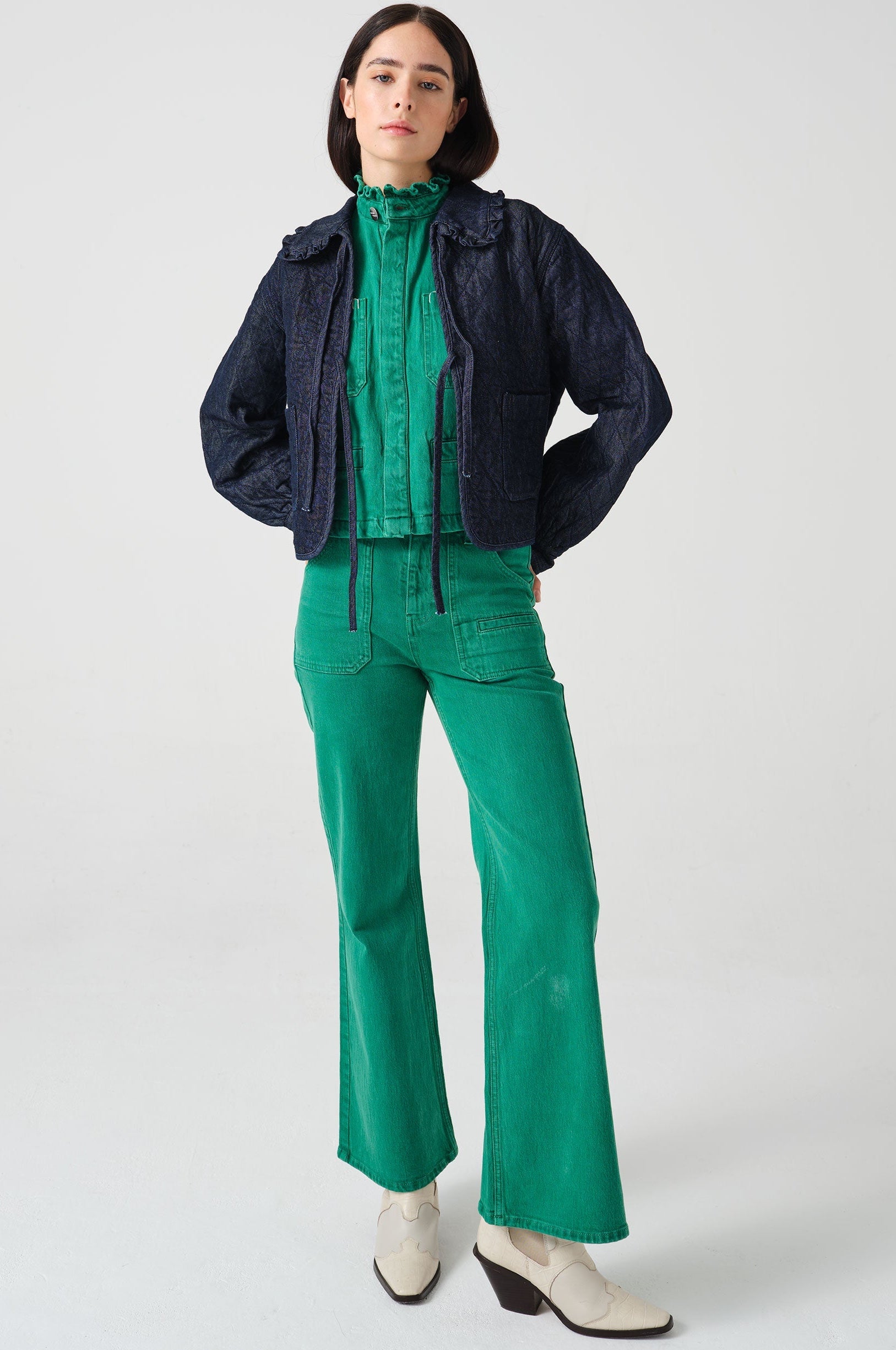 Woman wearing a dark blue denim quilted jacket with a slightly frilled collar and tie closures with a green denim shirt and green denim jeans