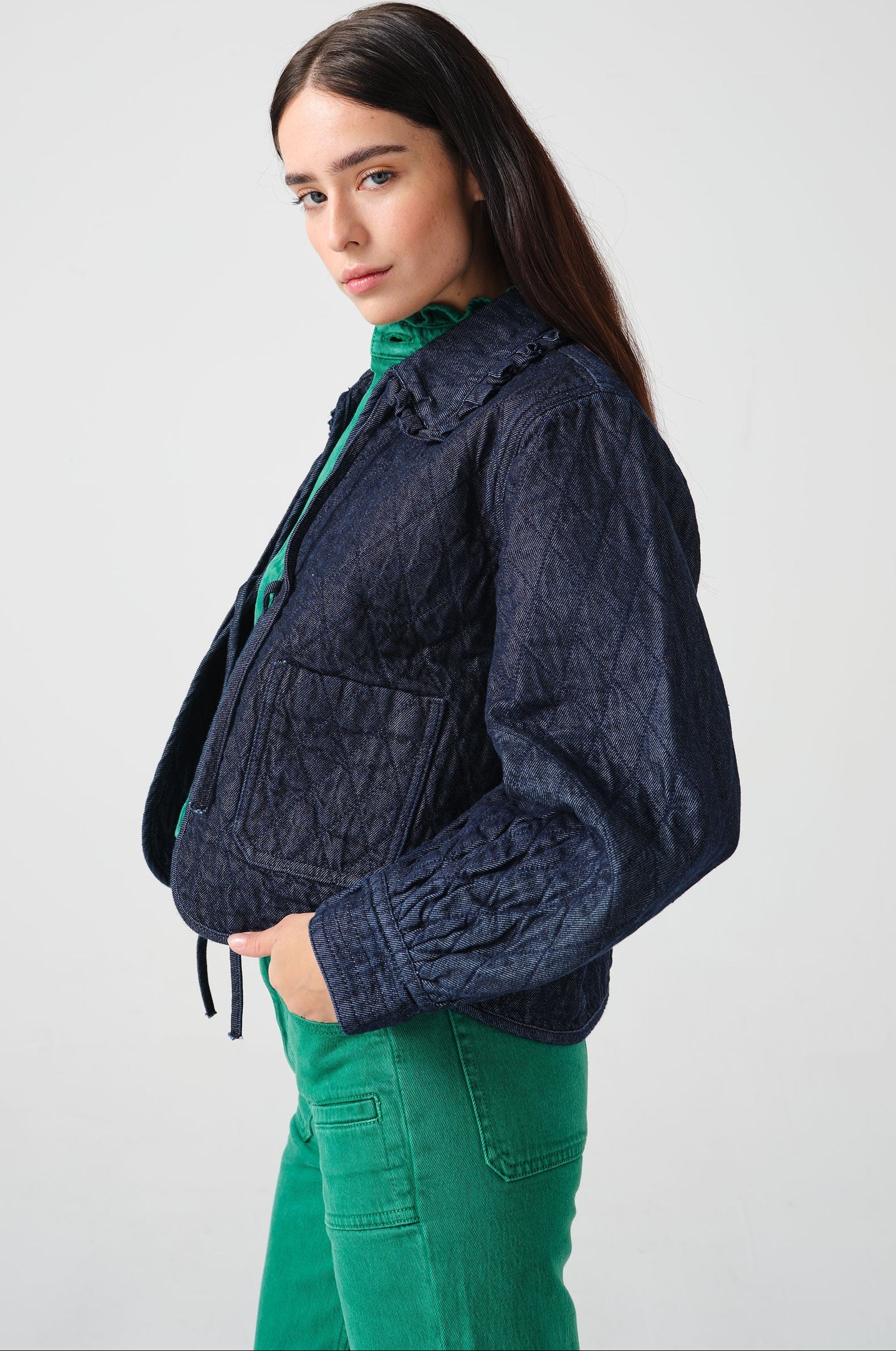 Woman wearing a dark blue denim quilted jacket with a slightly frilled collar and tie closures with a green denim shirt and green denim jeans