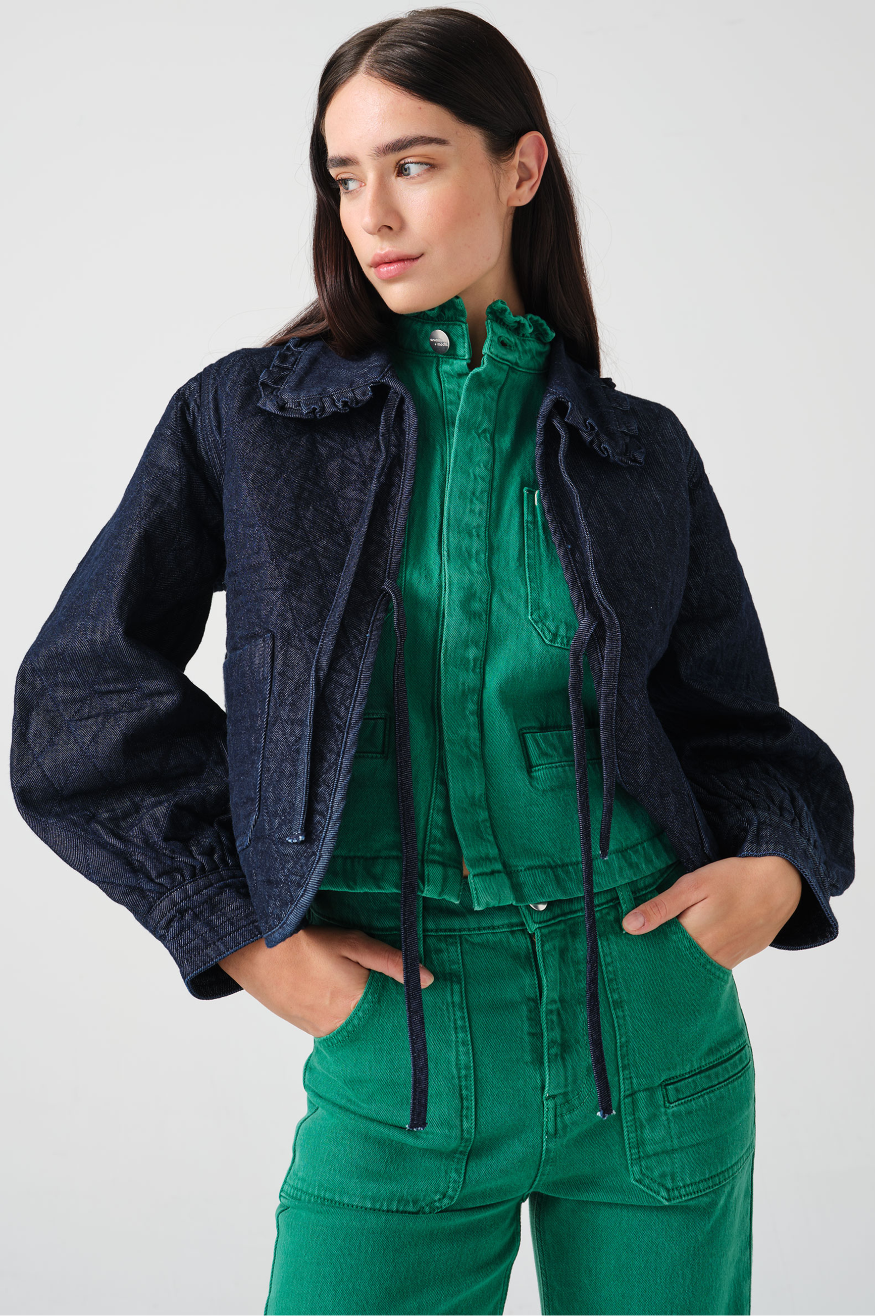 Woman wearing a dark blue denim quilted jacket with a slightly frilled collar and tie closures with a green denim shirt and green denim jeans