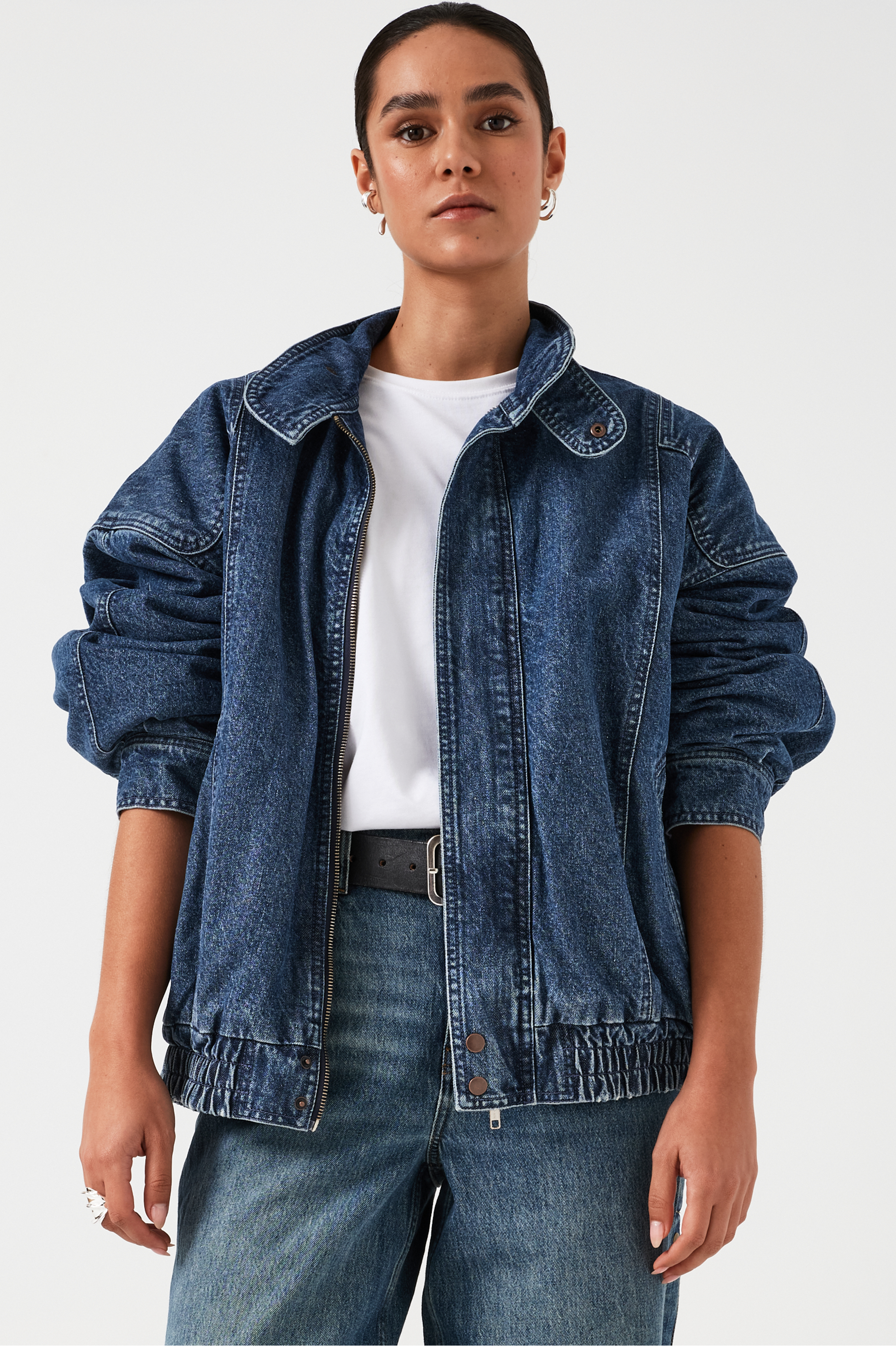 A woman wearing an oversized padded and seamed blue denim retro style bomber jacket with a round neckline, zip, and button closures at the top, bottom, and collar with a white top and blue jeans against a white backdrop