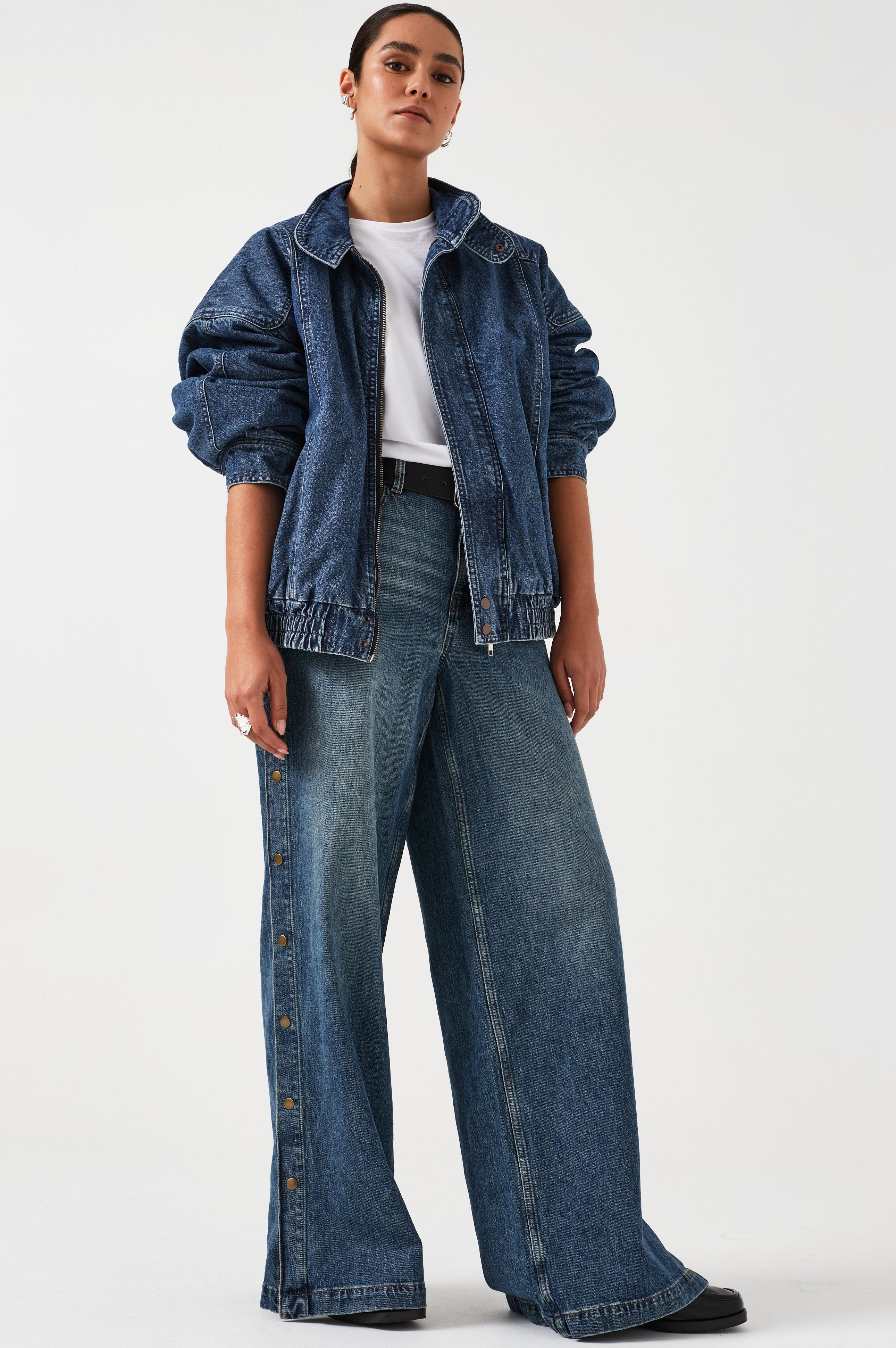 A woman wearing an oversized padded and seamed blue denim retro style bomber jacket with a round neckline, zip, and button closures at the top, bottom, and collar with a white top and blue jeans against a white backdrop