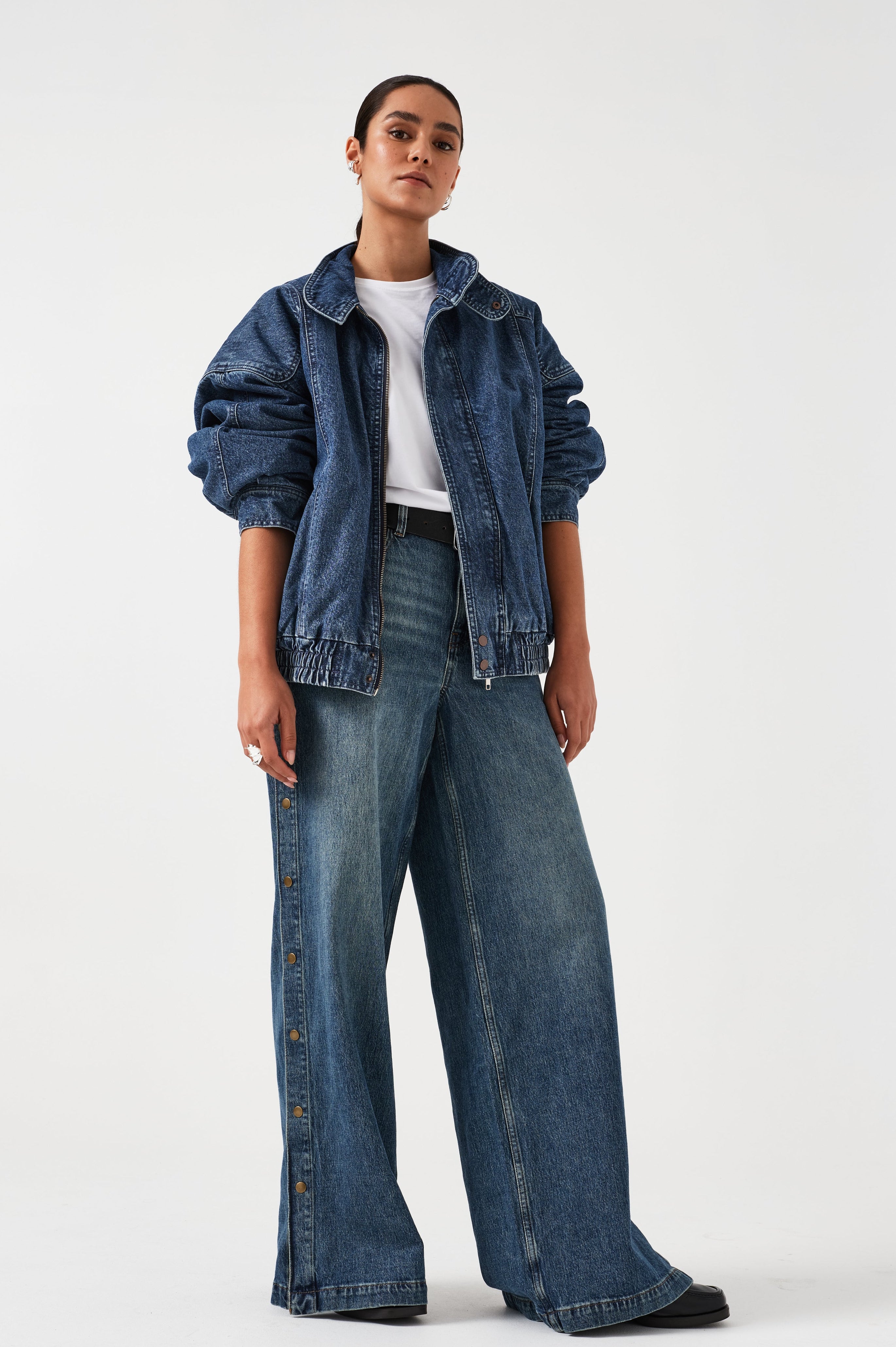 A woman wearing an oversized padded and seamed blue denim retro style bomber jacket with a round neckline, zip, and button closures at the top, bottom, and collar with a white top and blue jeans against a white backdrop