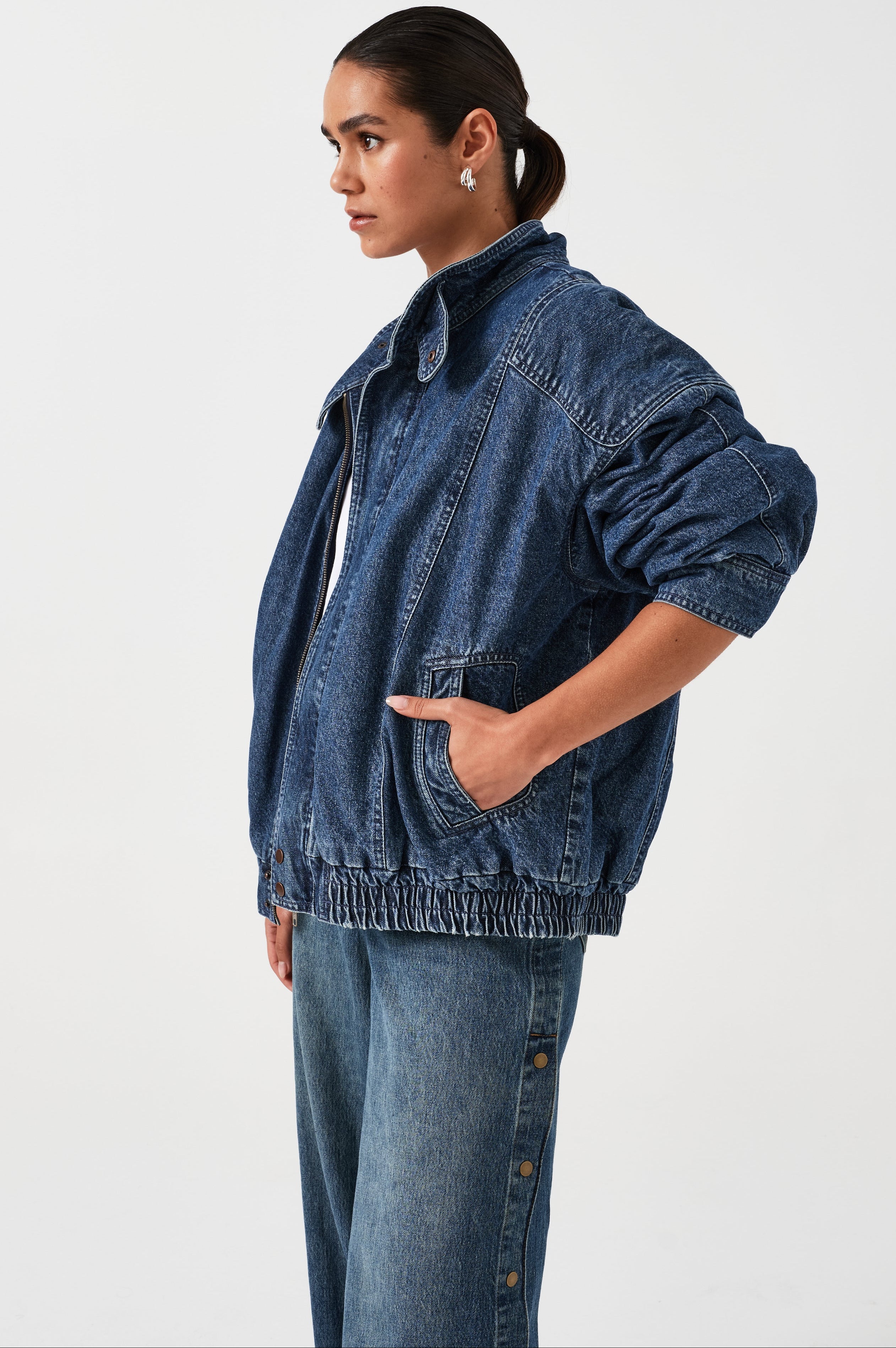 A woman wearing an oversized padded and seamed blue denim retro style bomber jacket with a round neckline, zip, and button closures at the top, bottom, and collar with a white top and blue jeans against a white backdrop