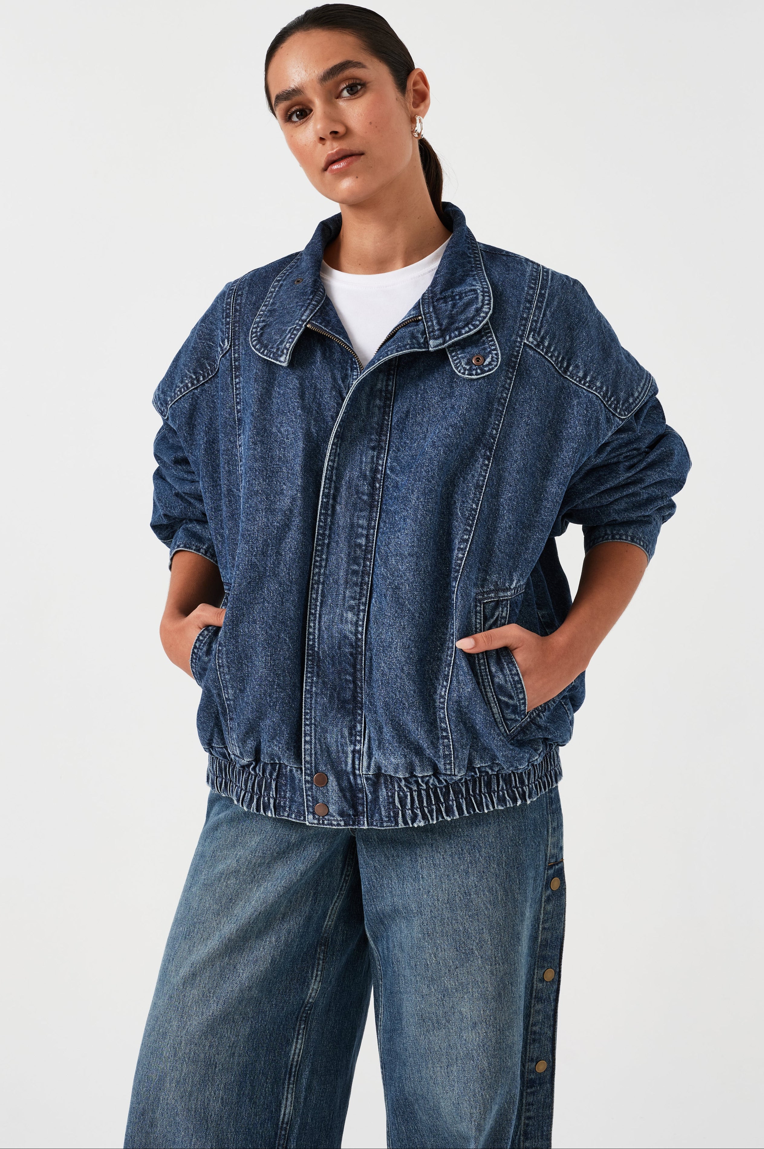 A woman wearing an oversized padded and seamed blue denim retro style bomber jacket with a round neckline, zip, and button closures at the top, bottom, and collar with a white top and blue jeans against a white backdrop