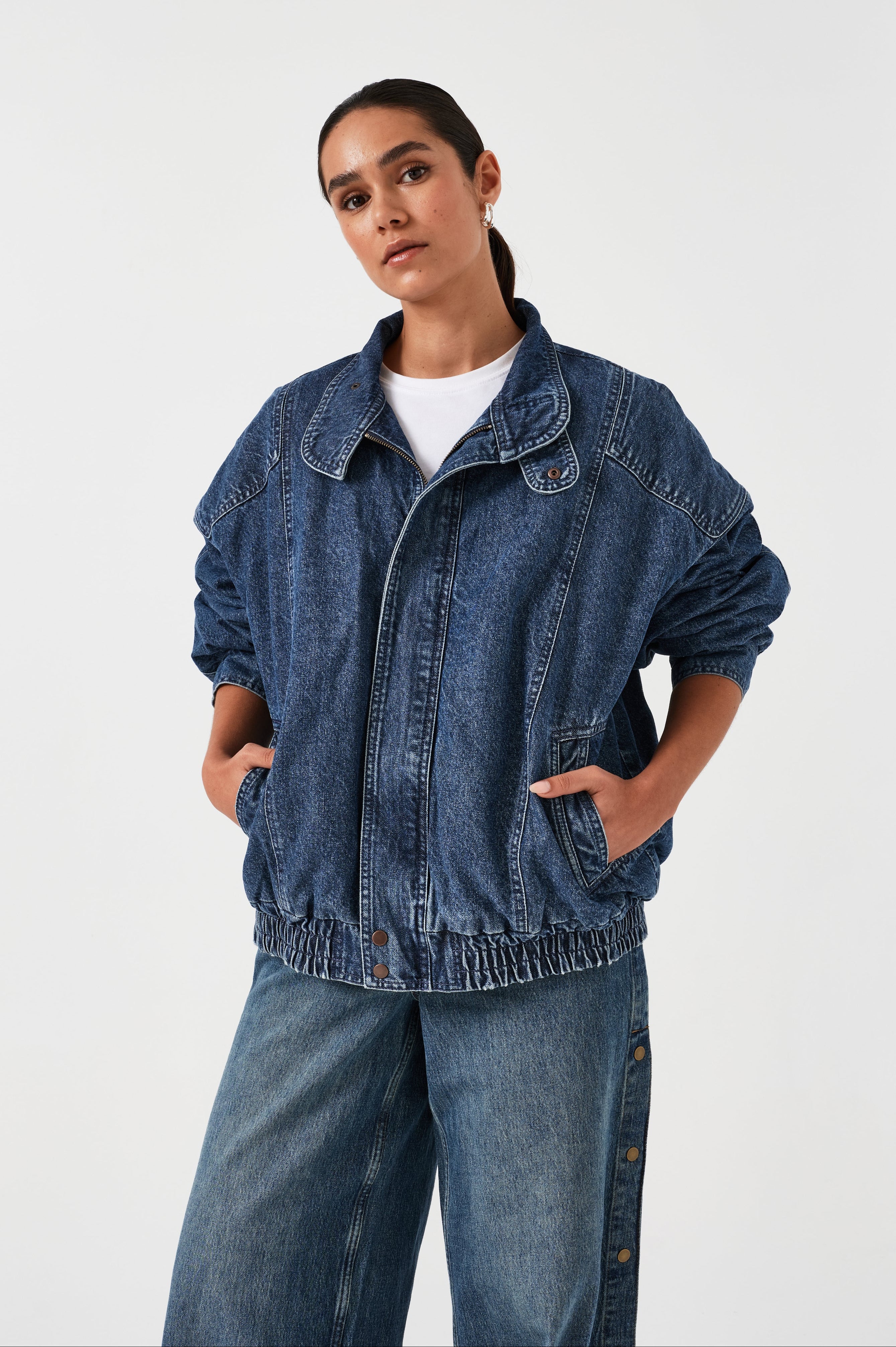 A woman wearing an oversized padded and seamed blue denim retro style bomber jacket with a round neckline, zip, and button closures at the top, bottom, and collar with a white top and blue jeans against a white backdrop