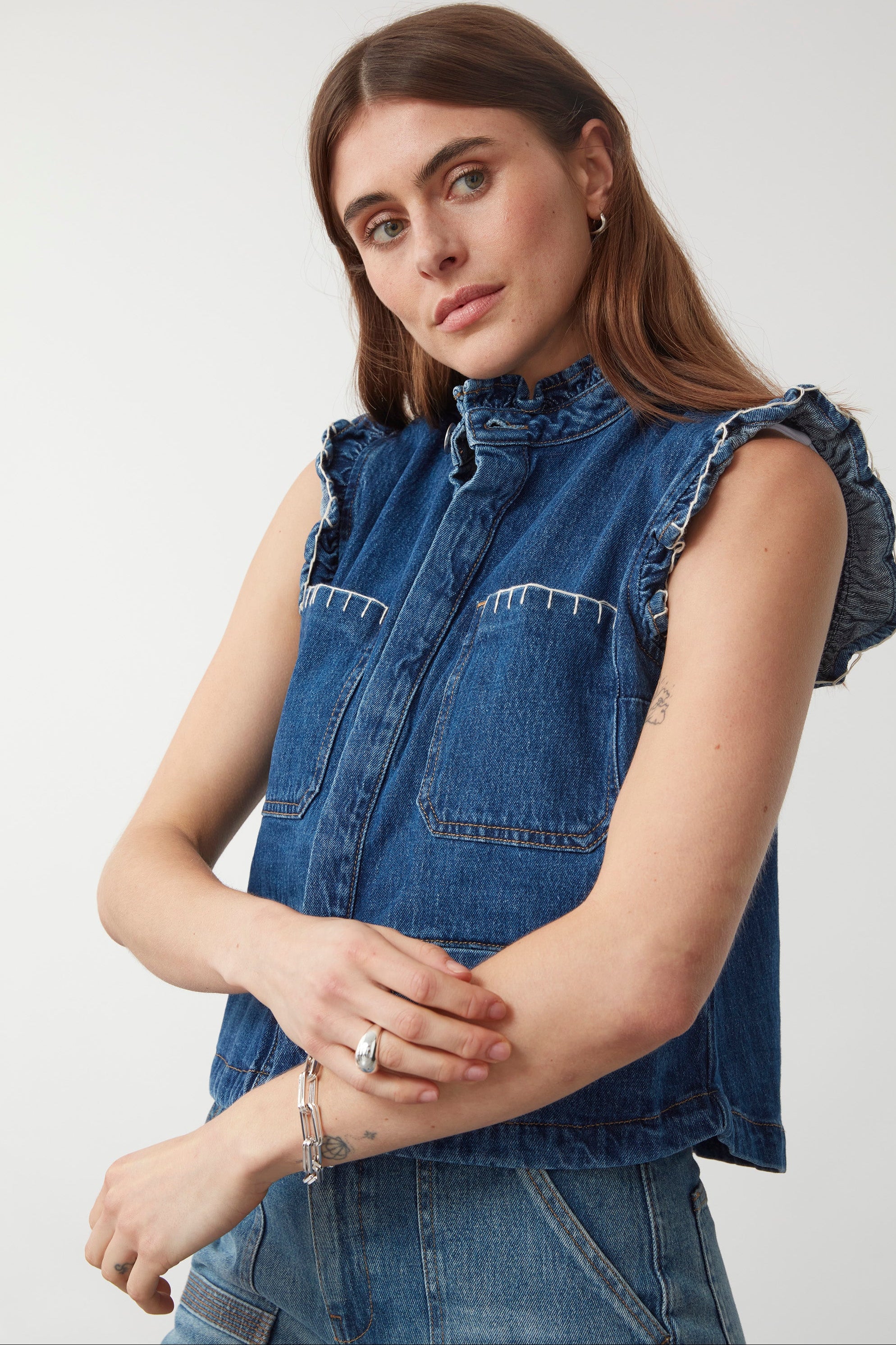 woman wearing a blue denim vest/ waistcoat with our signature white stitch detailing, two large patch pockets, and a frill collar with blue jeans against a white background