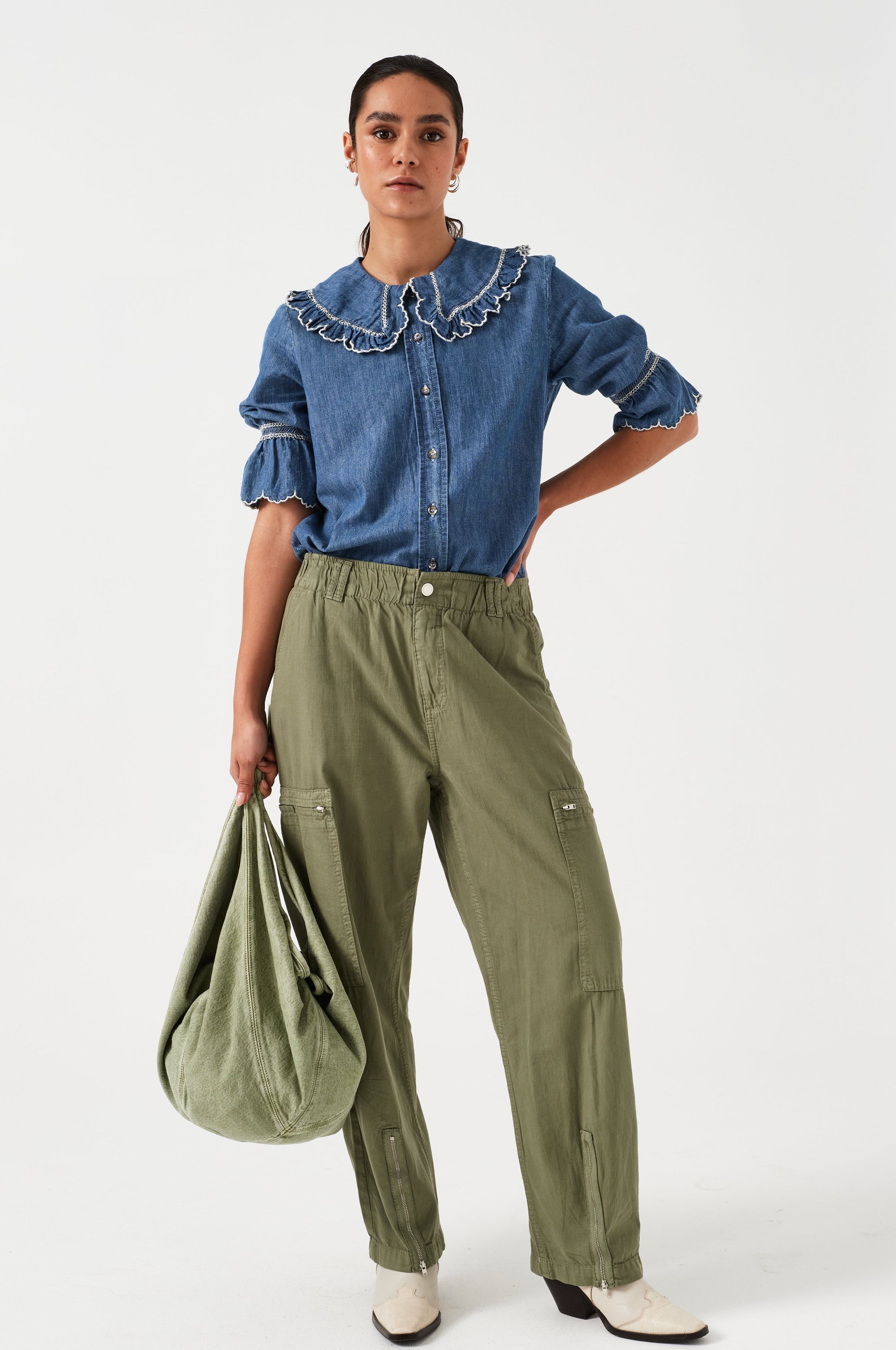 woman standing wearing lightweight cargo style khaki pants with deep zip pockets and an elastic waist and a denim blouse with a khaki bag on a white background