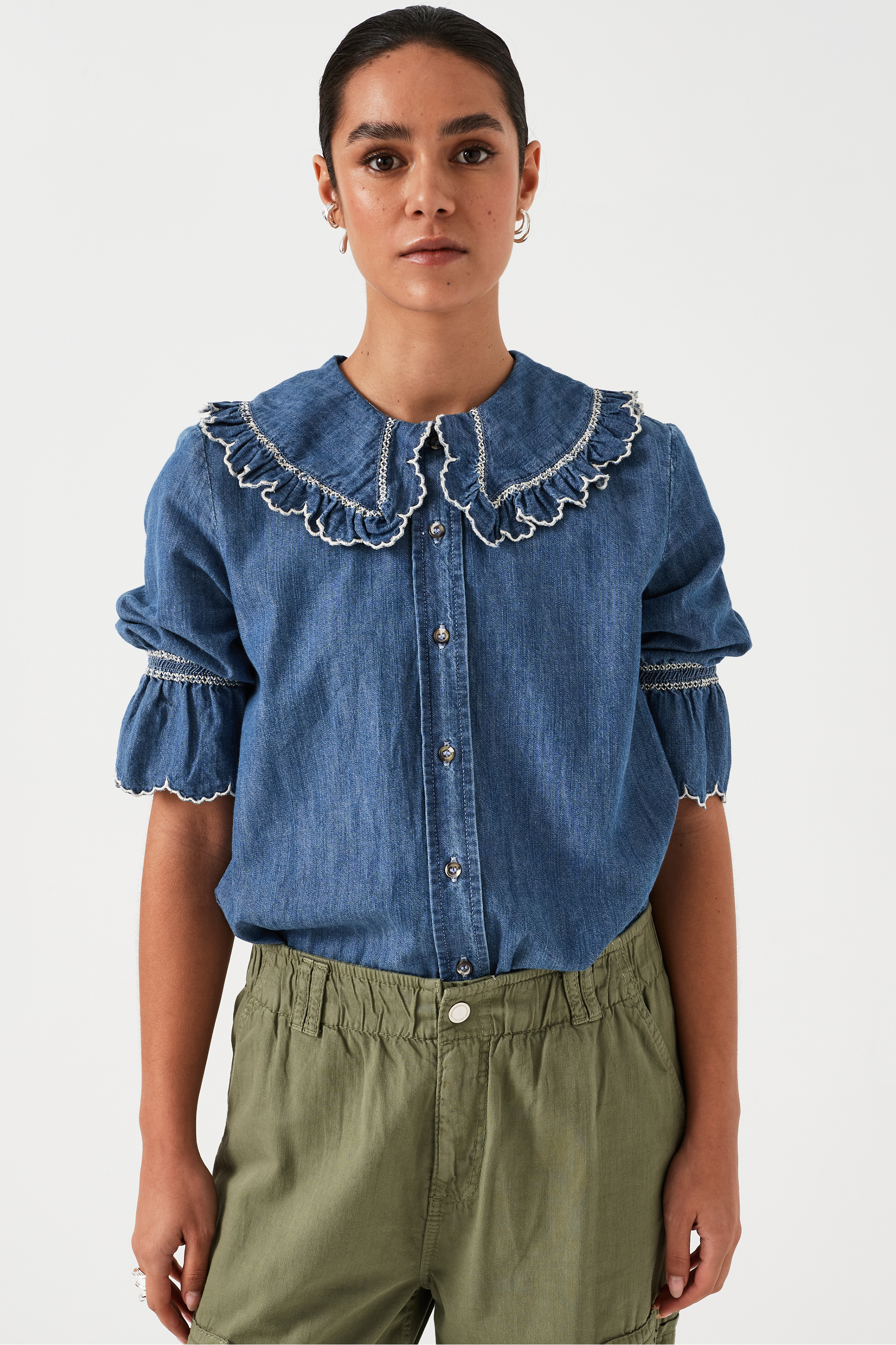 woman wearing an indigo denim blouse with white embroidery frill on the collar and sleeves and khaki pants on a white background