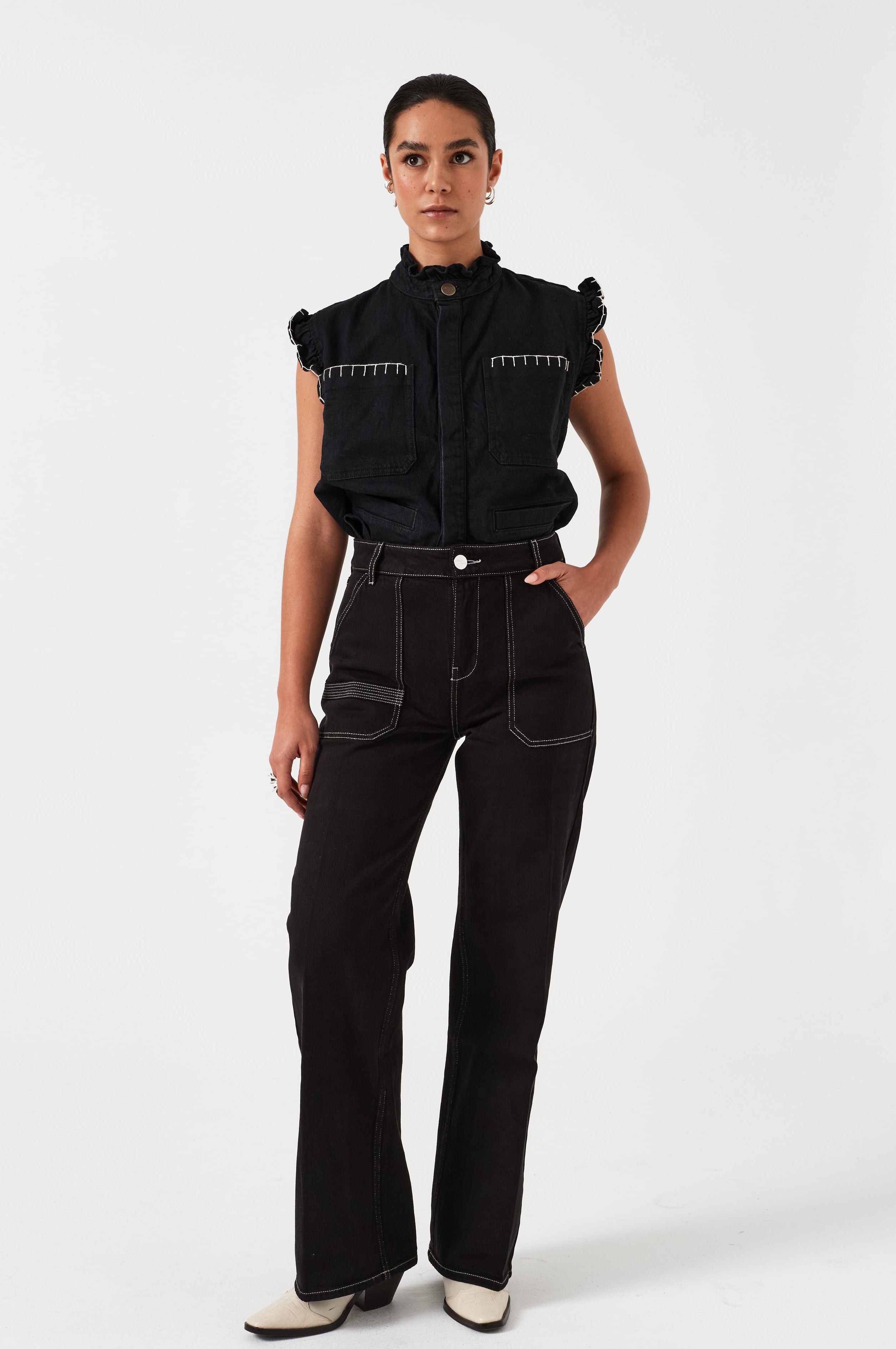 woman wearing a black vest/ waistcoat with a frill collar and our signature white stitching on two large patch pockets, paired with black jeans against a white backdrop