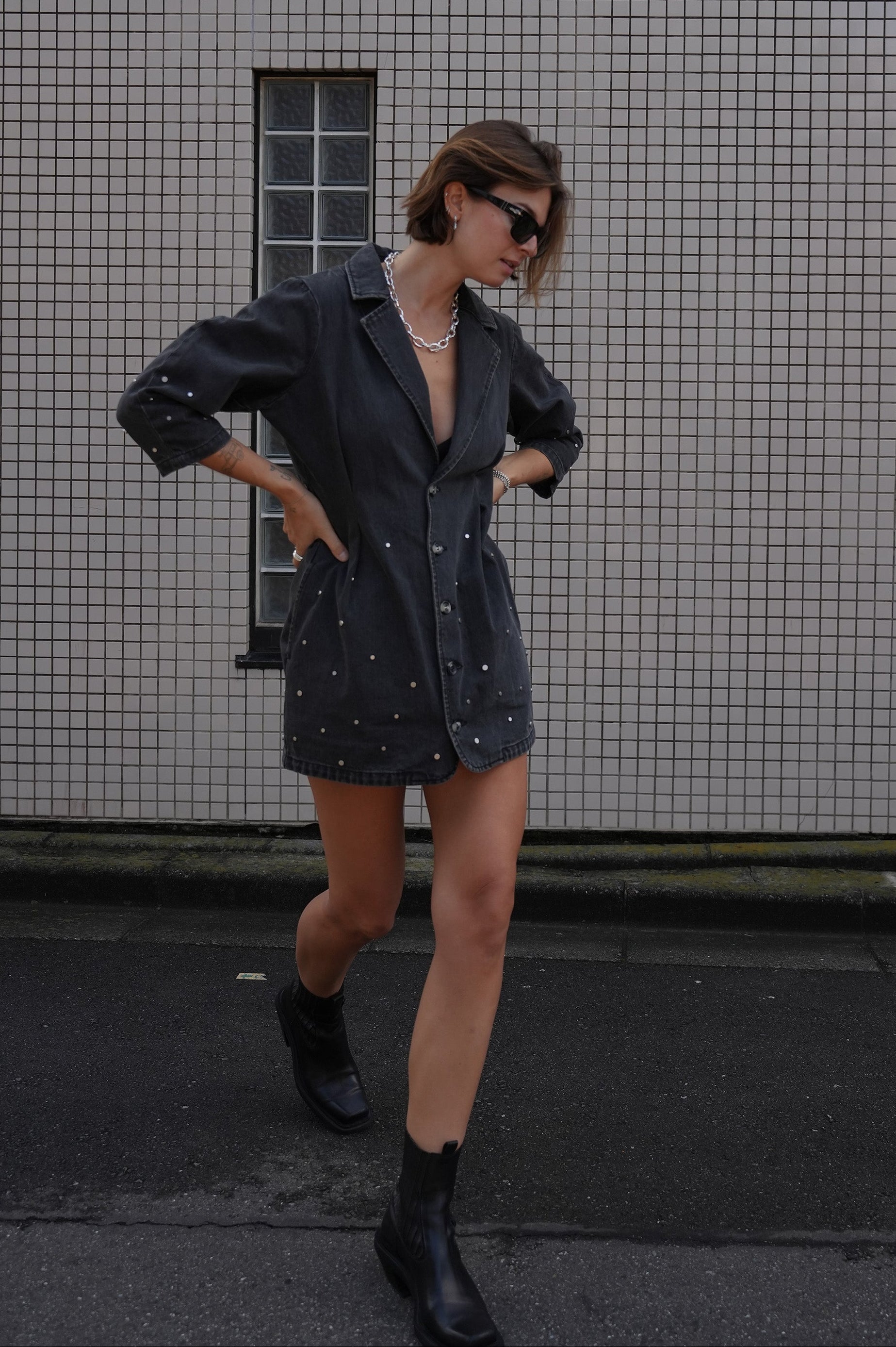 Woman wearing a black denim dress with silver studs (embellishments) and a tailored cinched waist against a tiled wall.