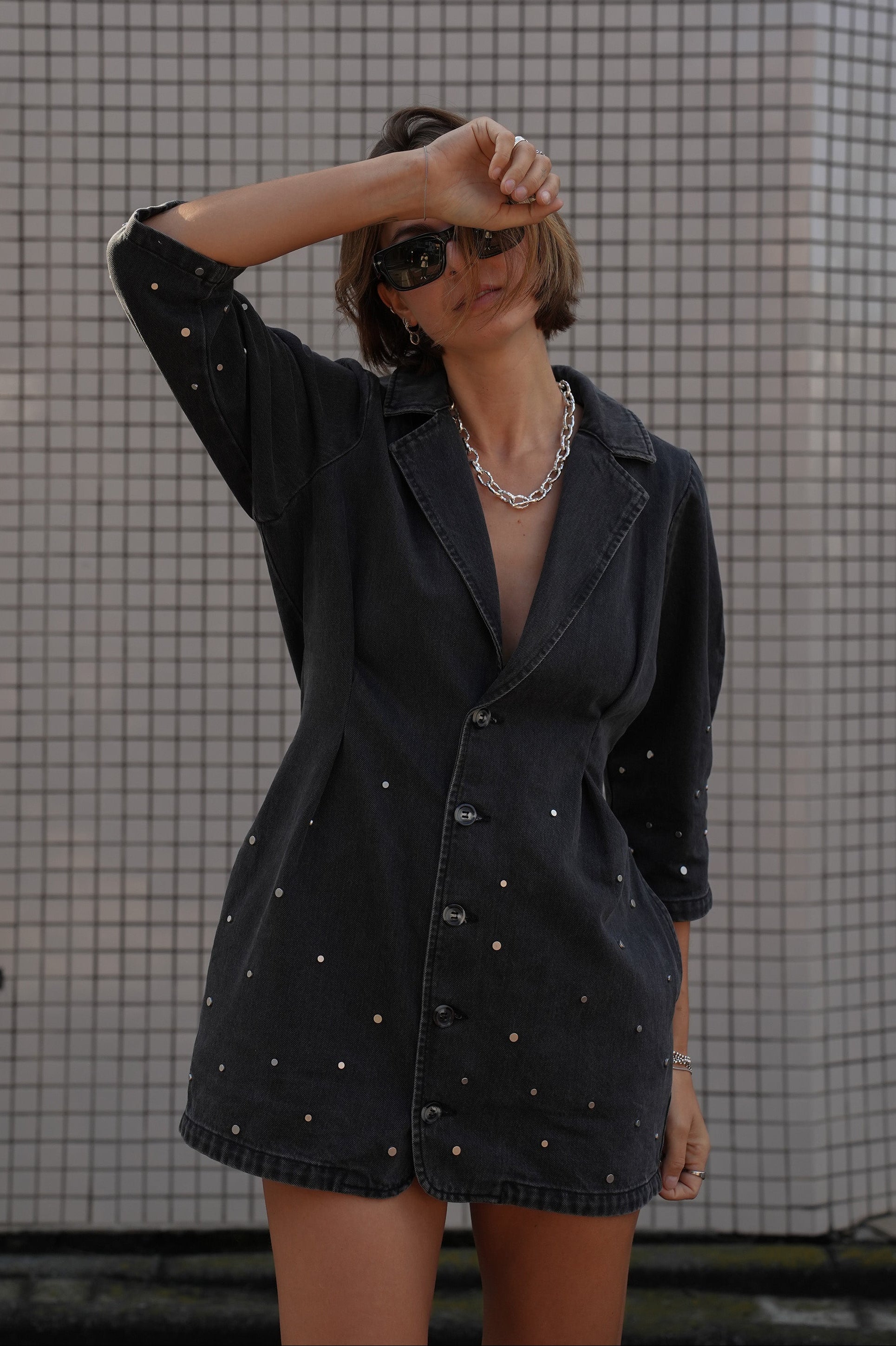 Woman wearing a black denim dress with silver studs (embellishments) and a tailored cinched waist against a tiled wall.