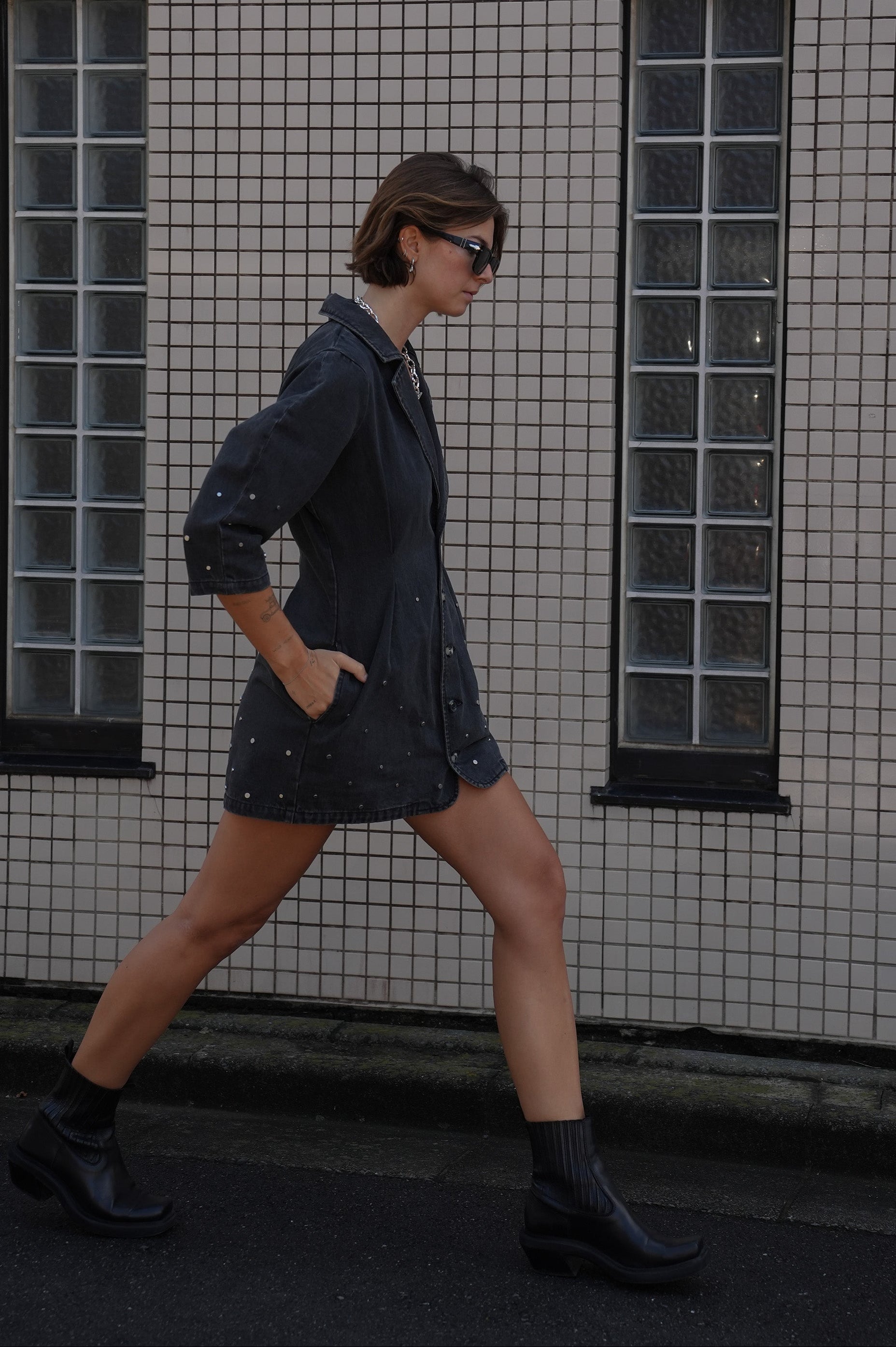 Woman wearing a black denim dress with silver studs (embellishments) and a tailored cinched waist against a tiled wall.