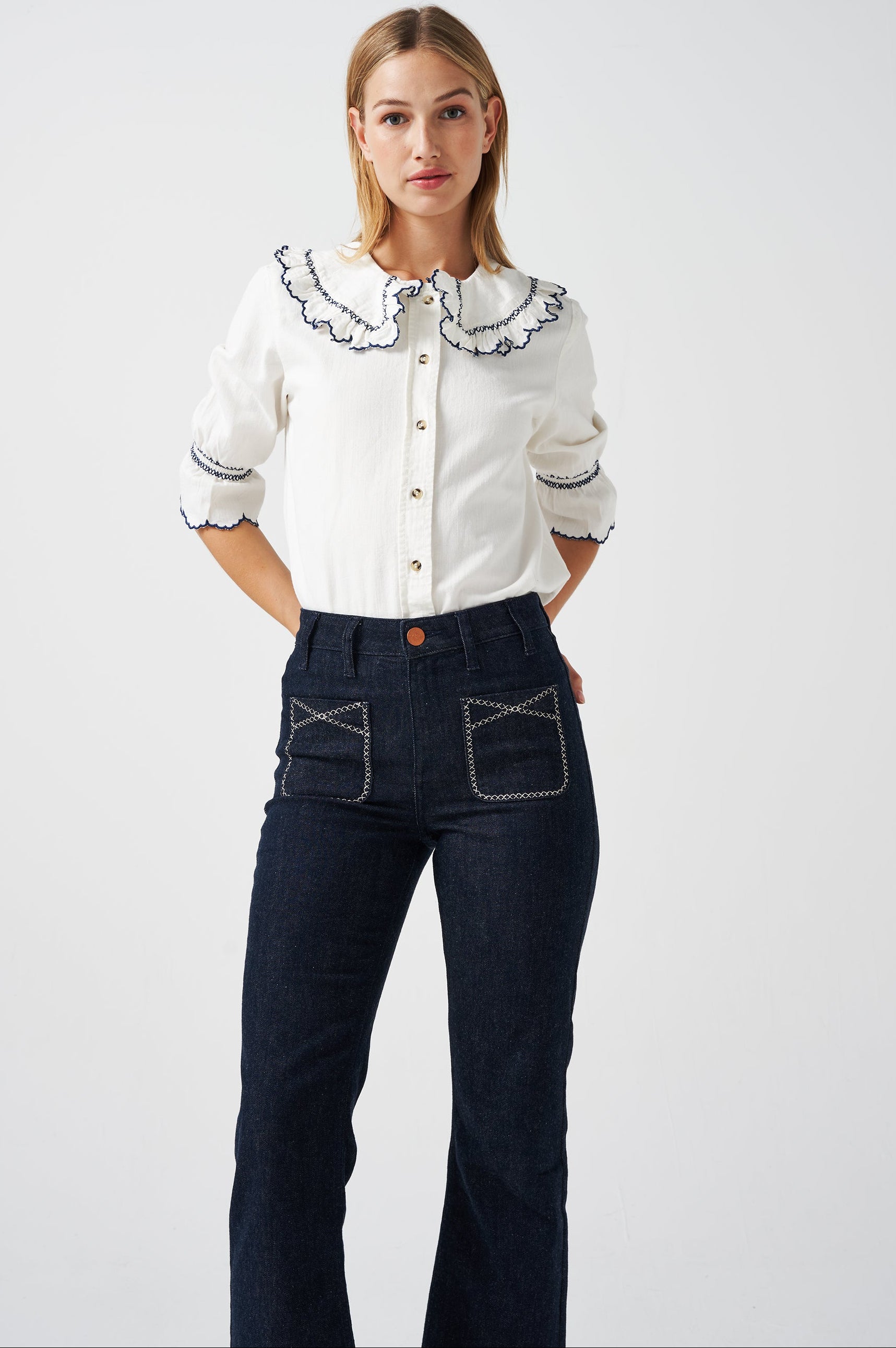 A woman wearing a white organic cotton blouse with dark blue embroiderery on frilled sleeves and a frill peek-a-boo collar with dark blue jeans on a white background