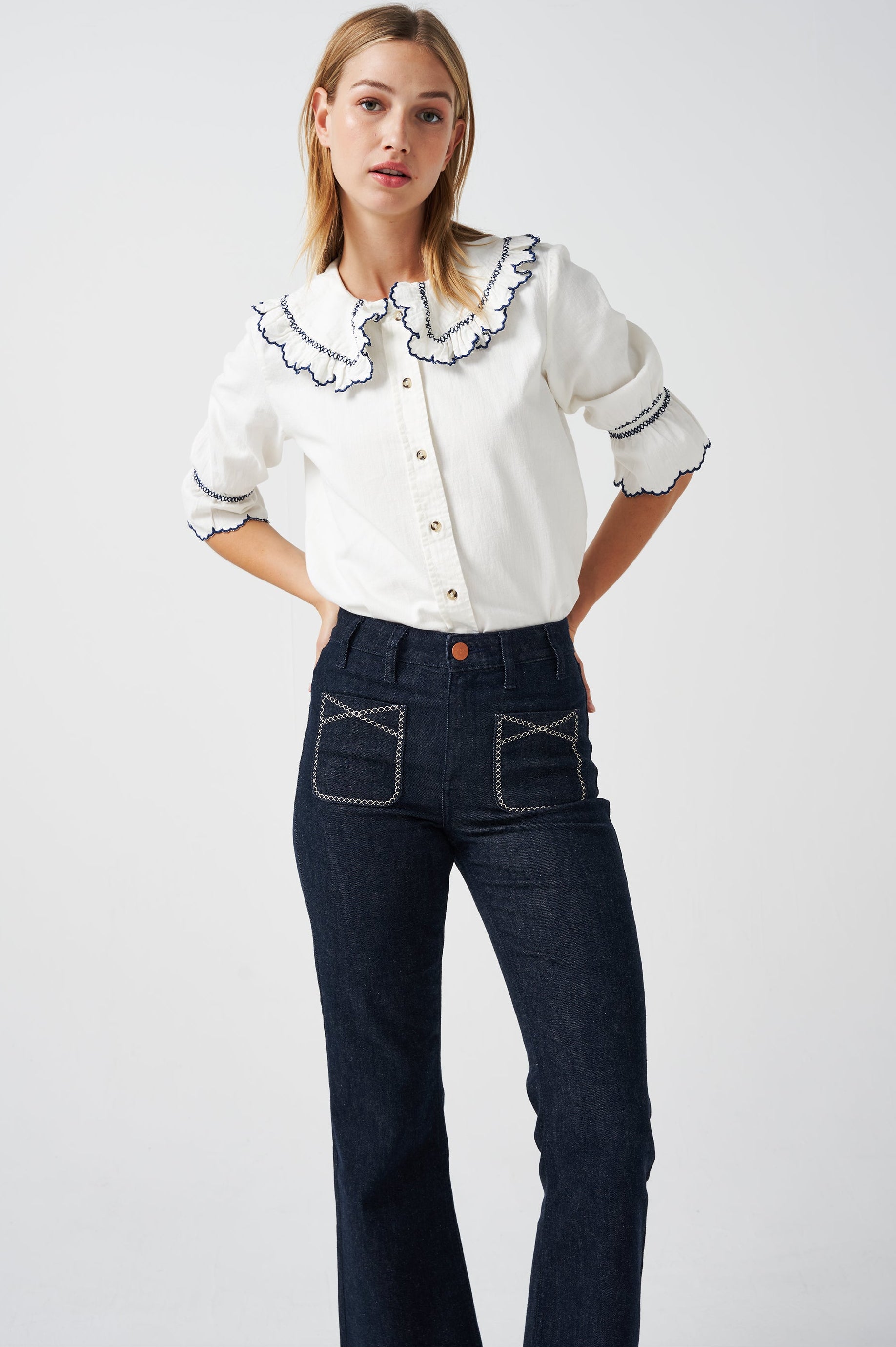 A woman wearing a white organic cotton blouse with dark blue embroiderery on frilled sleeves and a frill peek-a-boo collar with dark blue jeans on a white background