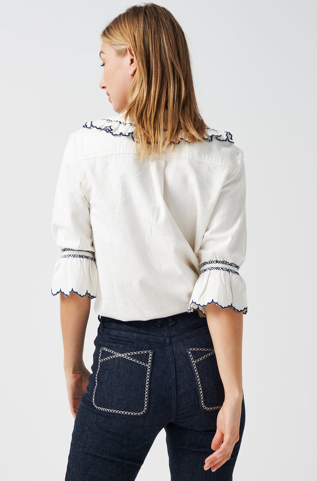 A woman wearing a white organic cotton blouse with dark blue embroiderery on frilled sleeves and a frill peek-a-boo collar with dark blue jeans on a white background