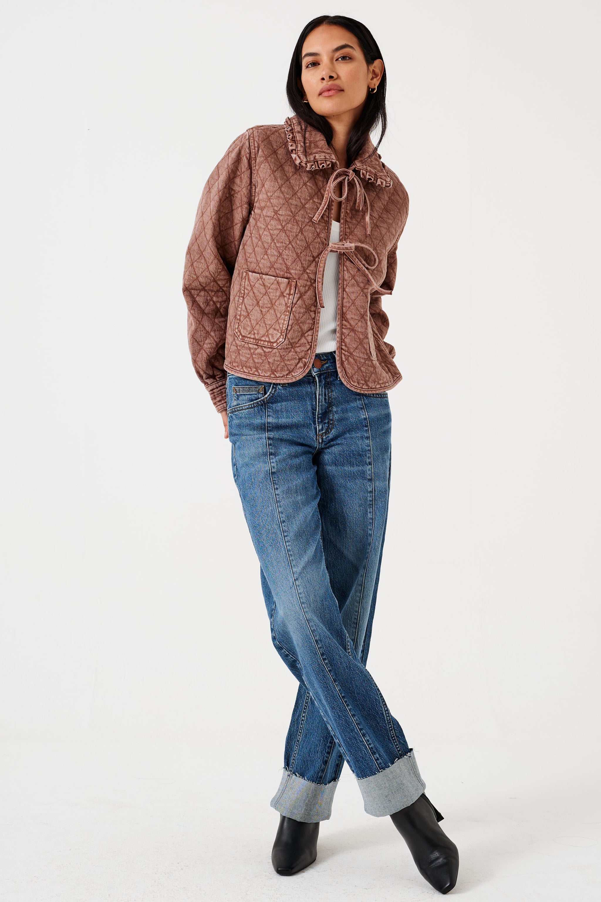 Woman wearing a brown quilted denim jacket with large pockets, a frill collar,  and front tie closure, paired with a blue jeans on a white background
