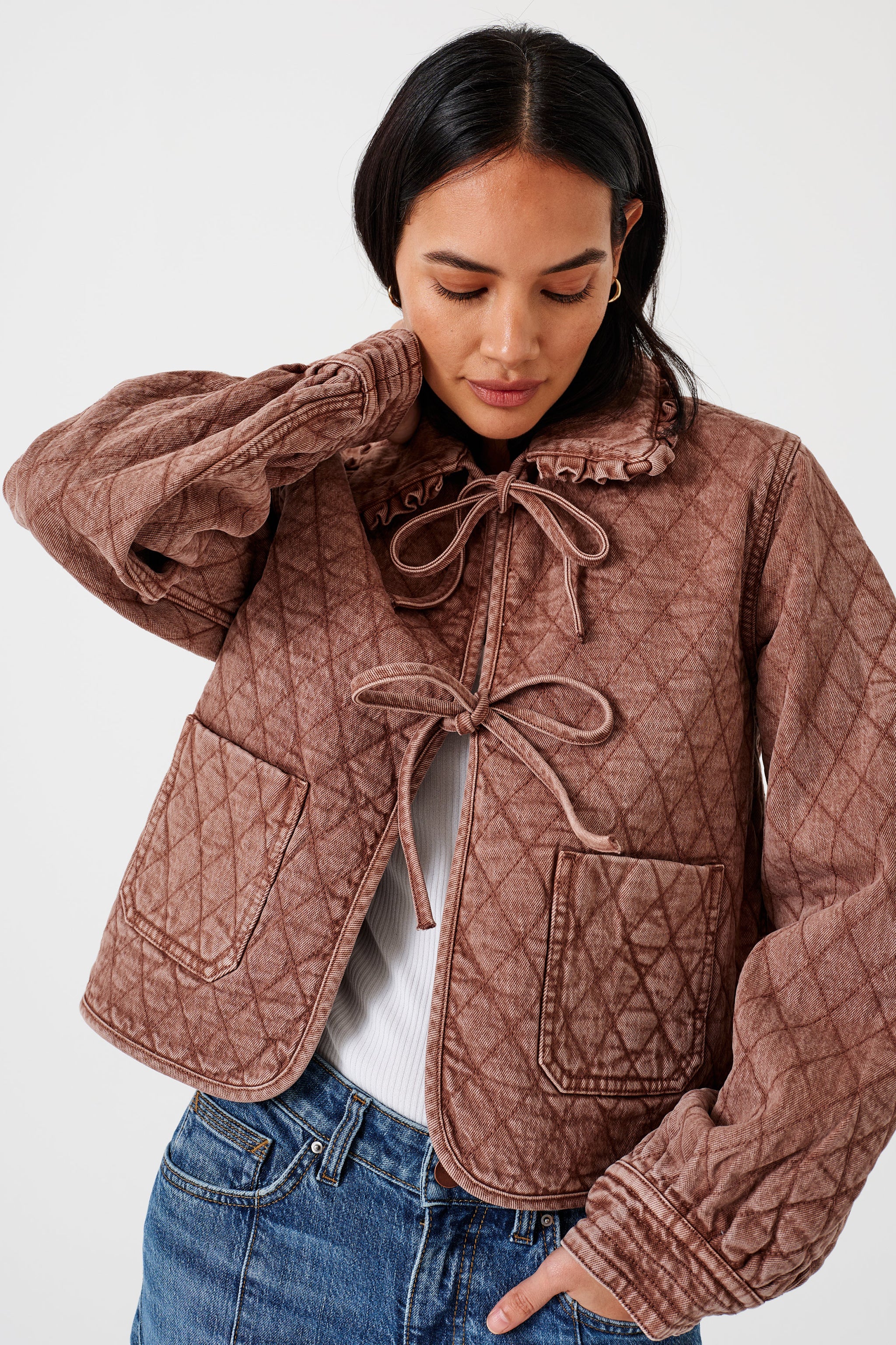 Woman wearing a brown quilted denim jacket with large pockets, a frill collar,  and front tie closure, paired with a blue jeans on a white background