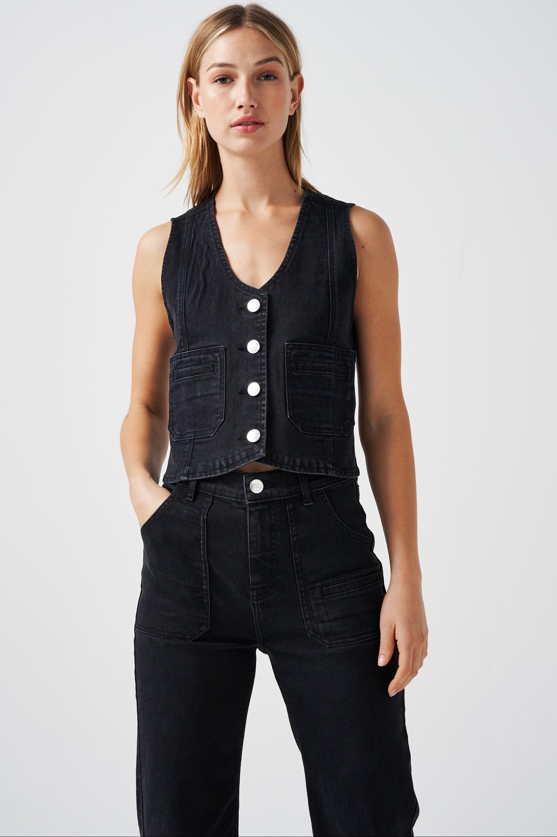 woman wearing a black denim waistcoat/ vest with two large patch pockets and black jeans on a white background
