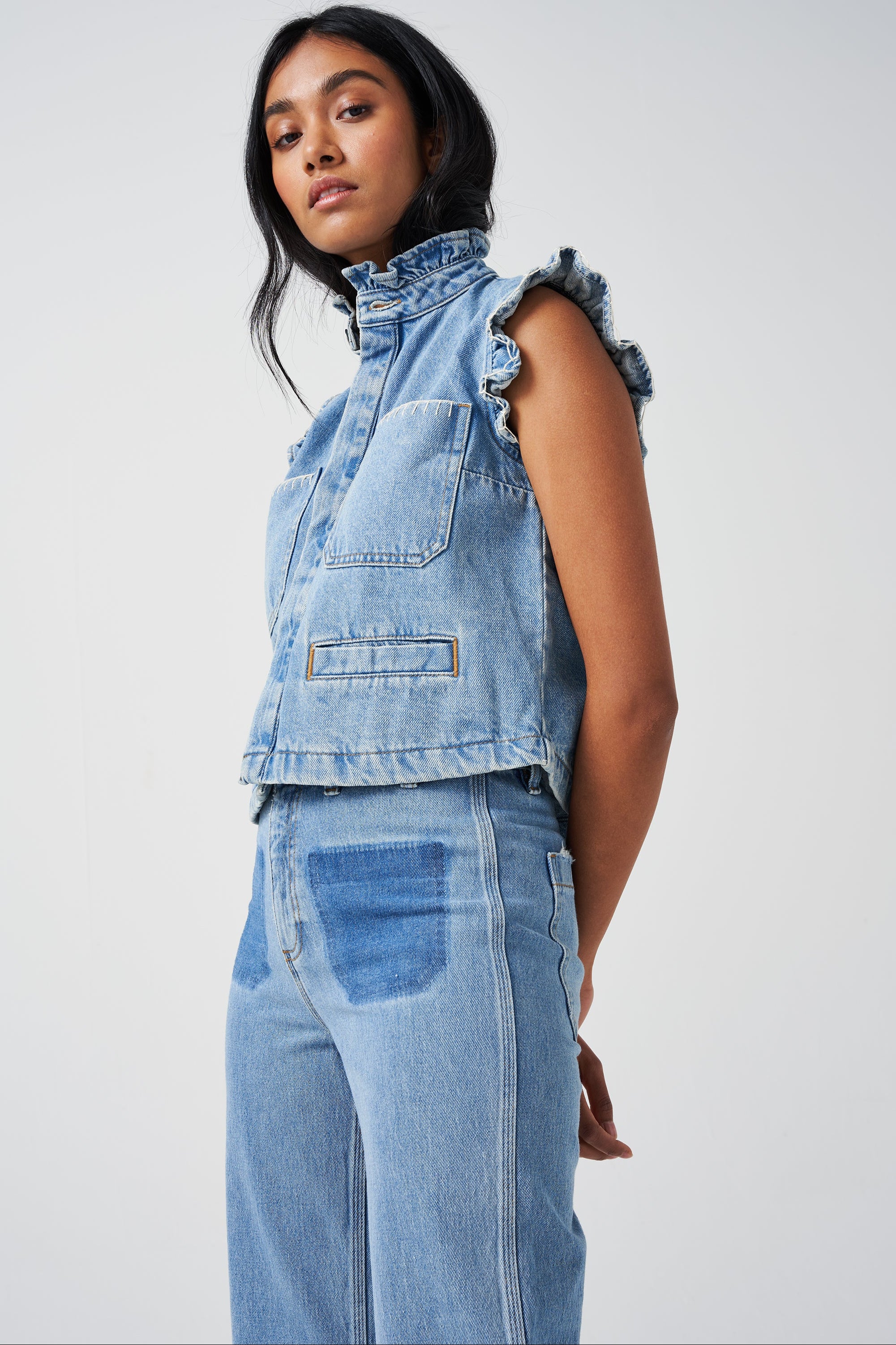 woman wearing a blue denim waistcoat/ vest with frill collar and sleeves, and two large patch pockets with white stitching, paired with blue jeans on a white background
