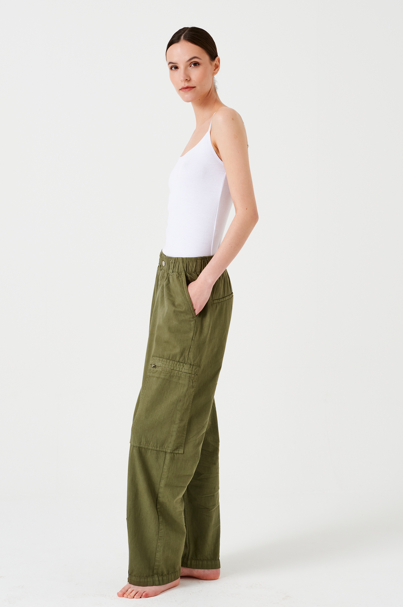 woman standing wearing lightweight cargo style khaki pants with deep zip pockets and an elastic waist and a white tank top on a white background.