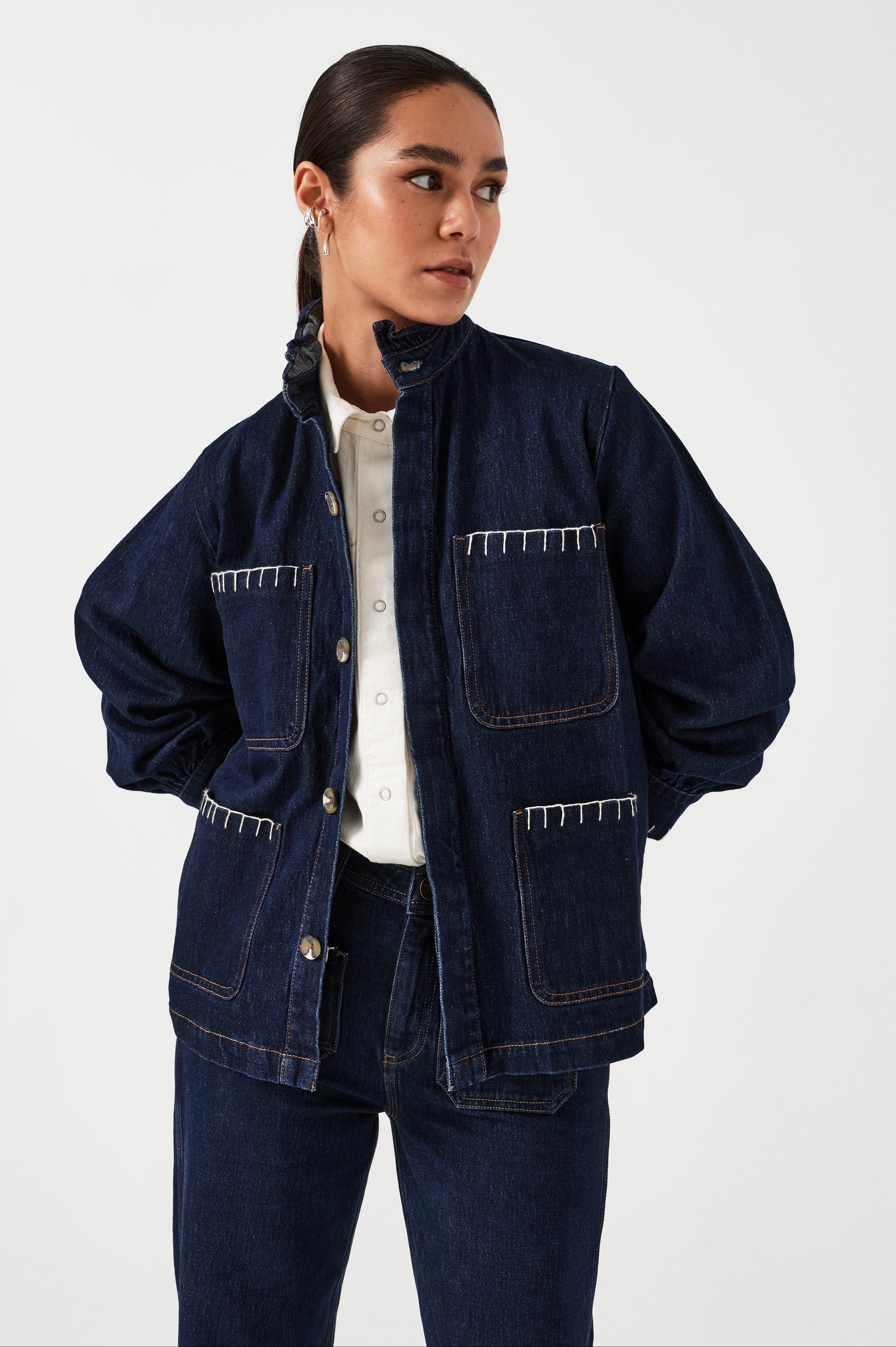 A woman wearing a dark blue vintage style jacket with a frill collar and our signature white blanket stitching on four large patch pockets, paired with a dark blue jeans and a white top against a white background