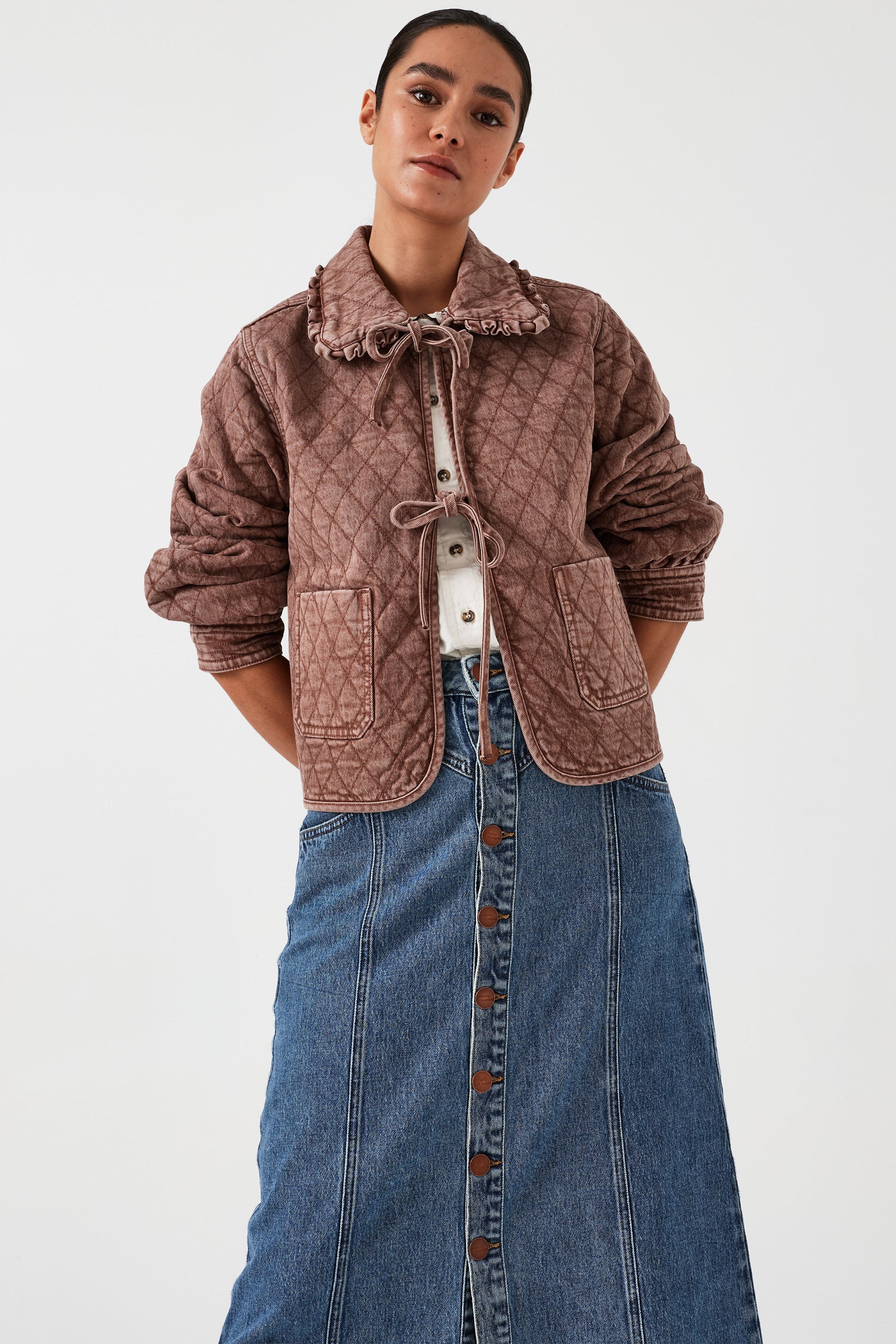 Woman wearing a brown quilted denim jacket with a frill collar and front tie closure, paired with a blue skirt on a white background